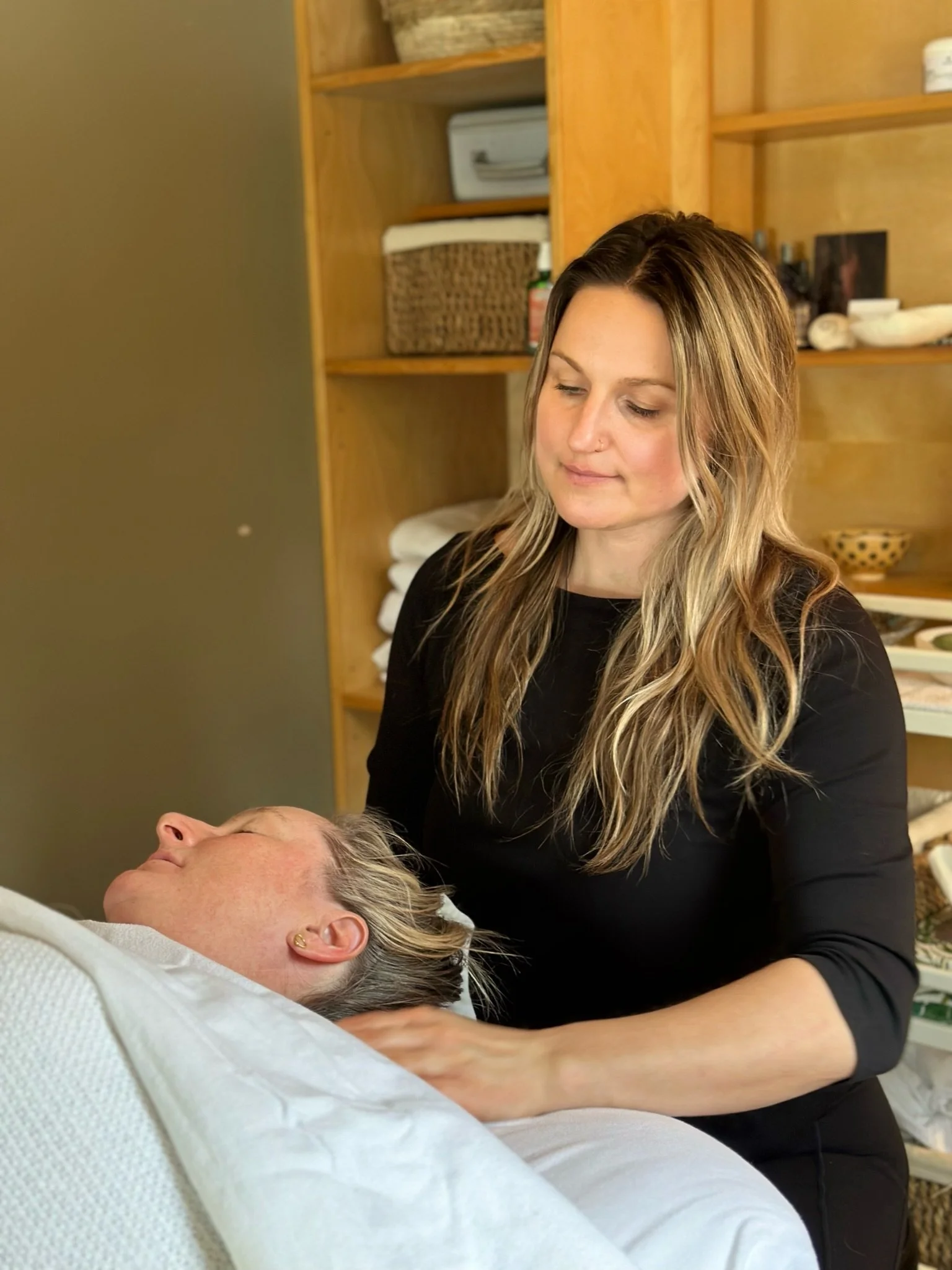 A woman giving a neck and shoulder massage to a person lying in a bed, in a room with wooden shelves.