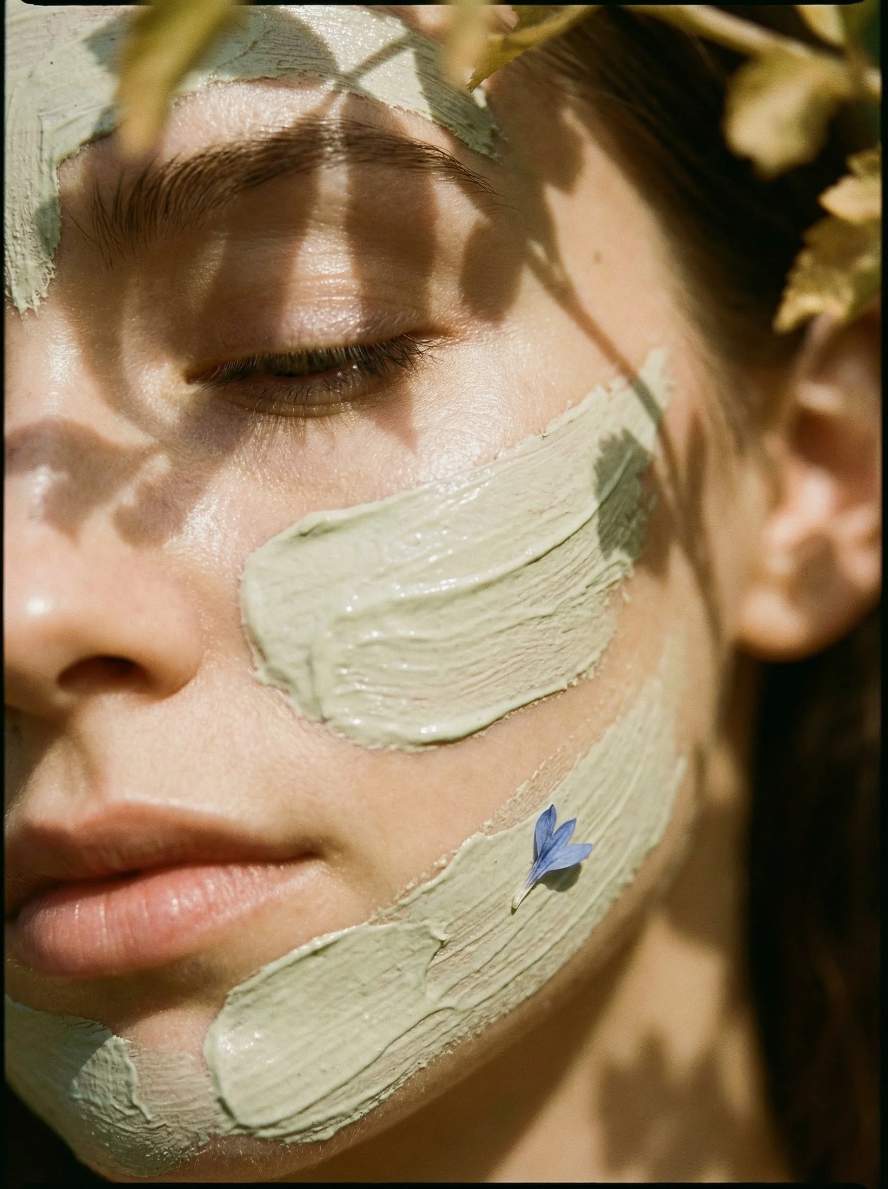 Close-up of a woman with her eyes closed, lying outdoors with green leaves casting shadows on her face. She has a clay facial mask applied on her cheeks and chin, with a small blue flower resting on her chin, suggesting a skincare or spa treatment.
