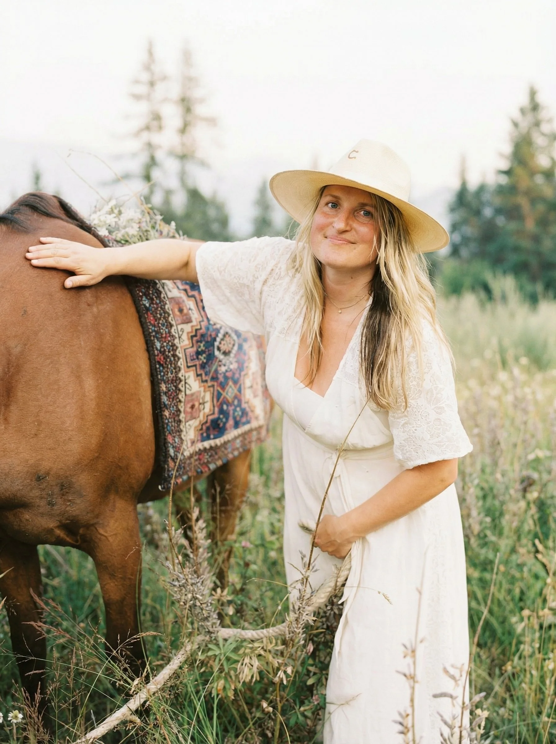 A woman with long blonde hair wearing a white dress and wide-brimmed hat, standing in a field with tall grasses and wildflowers, resting her hand on a brown horse that has a patterned saddle blanket.