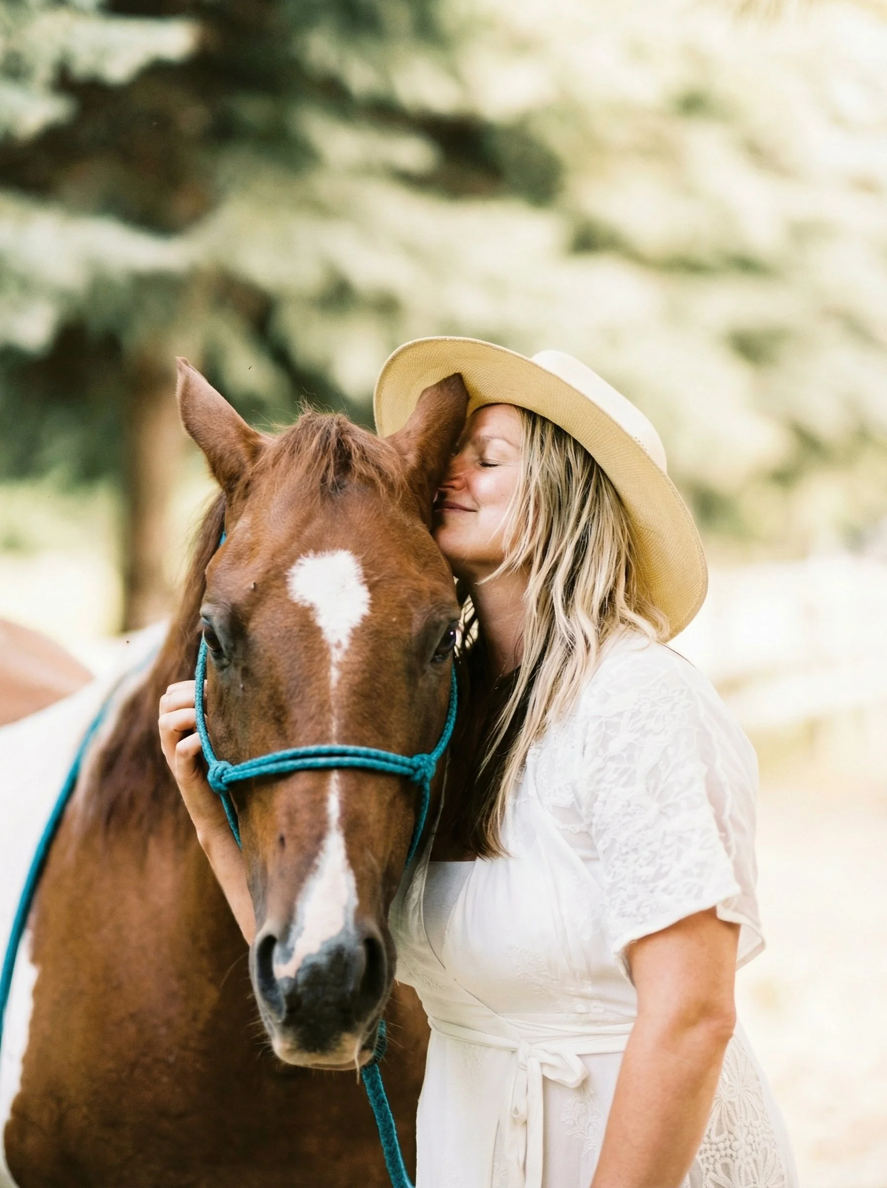 A woman in a white dress and a wide-brimmed hat hugging and kissing a brown horse with a white mark on its face, outdoors with blurred trees in the background.