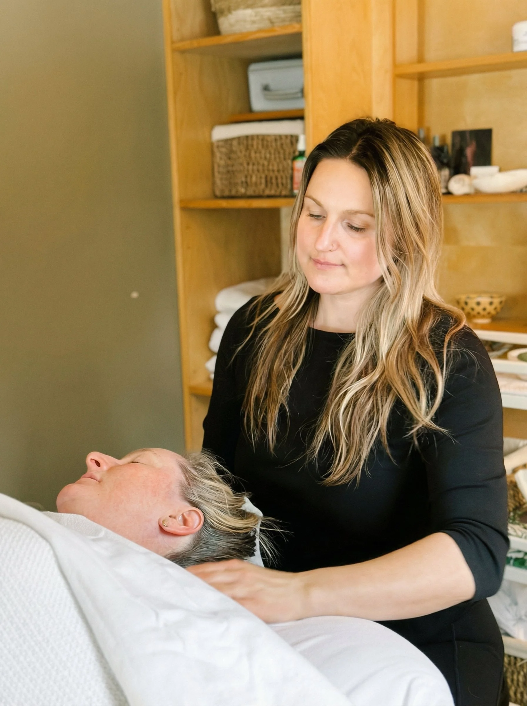 A woman with long, wavy blond hair gently cares for an elderly woman lying in bed, with a wooden shelf filled with folded towels and various items in the background.