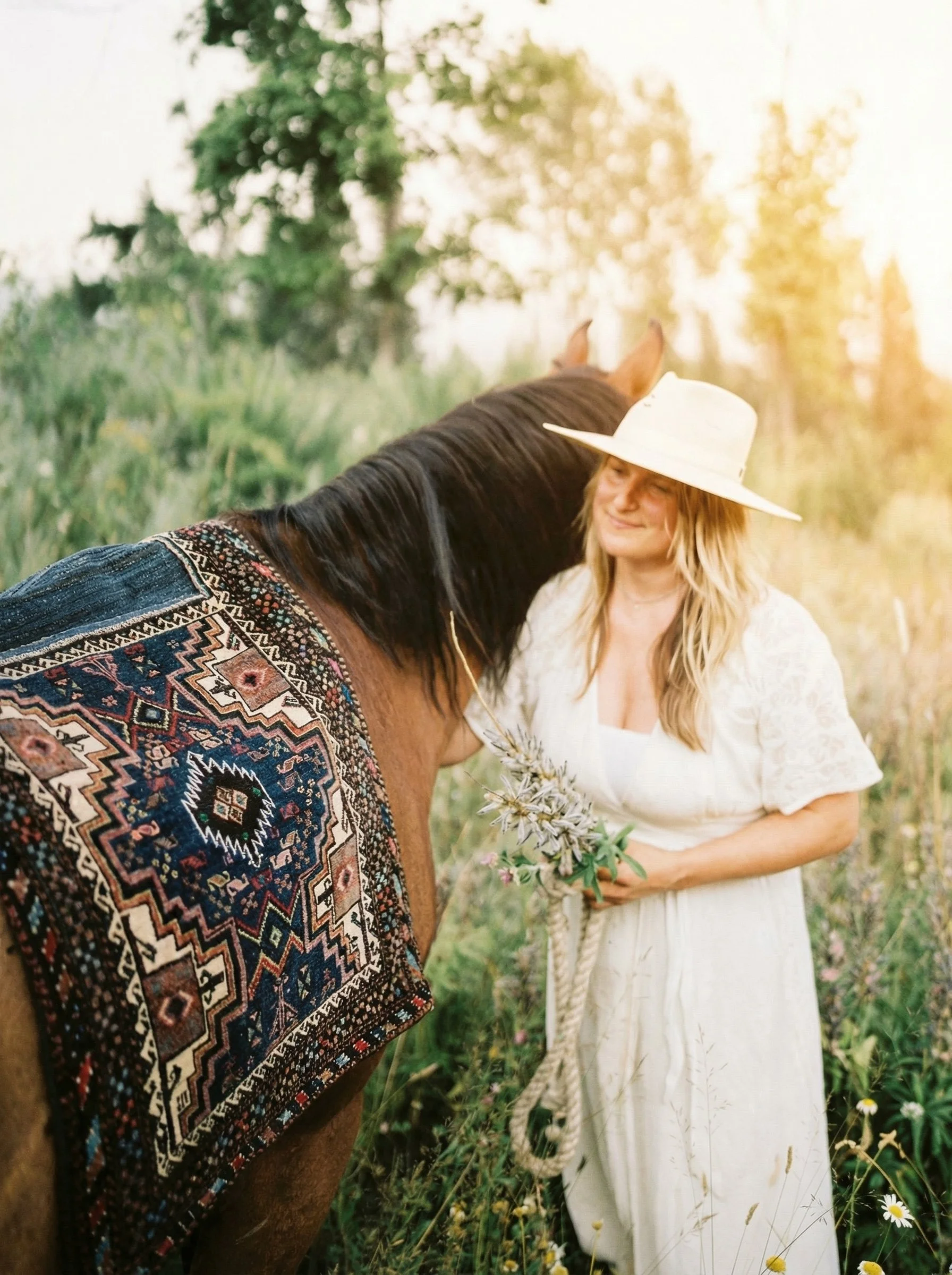 A woman in a white dress and large white hat is standing in a field of grass and wildflowers, gently holding a bouquet of flowers, while a horse with a decorated blanket is nuzzling her face.