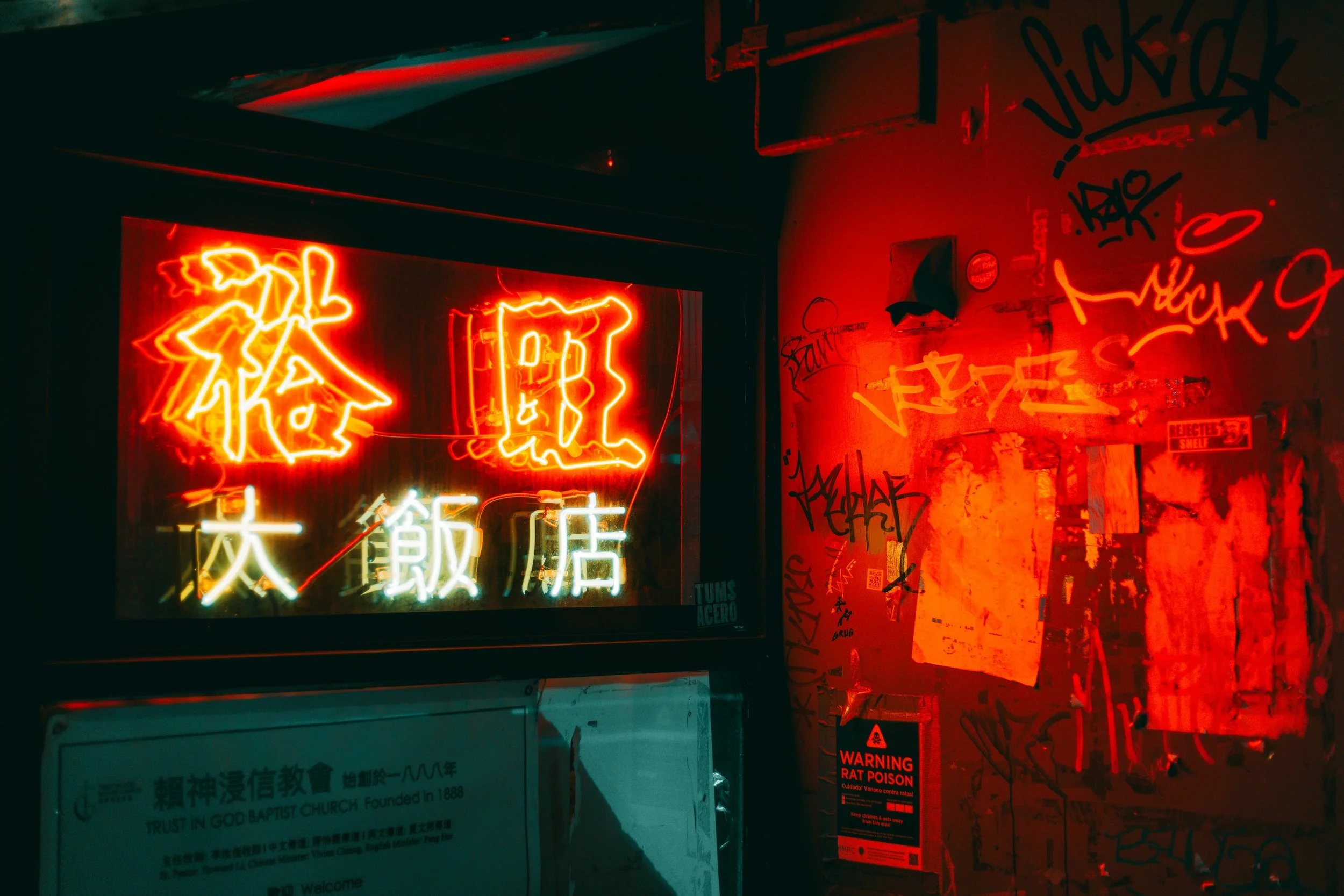 Nighttime scene with red neon signs in Chinese characters reflected on a glass surface, graffiti-covered wall, and a warning sign about rat poison.