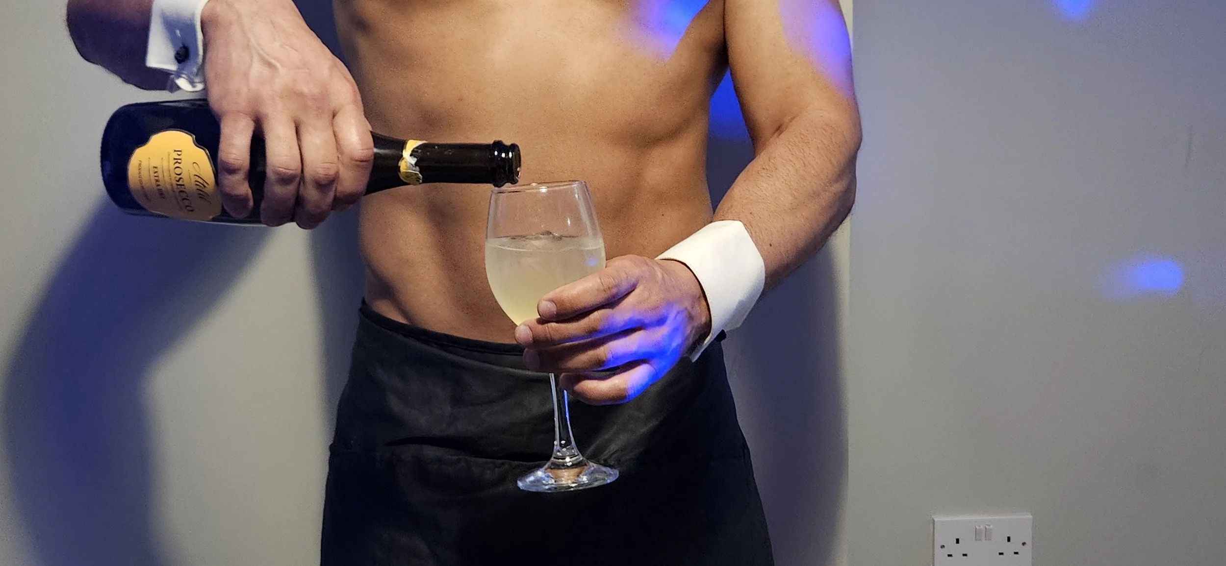 A shirtless man wearing black trousers and a white cuff shirt is pouring champagne into a wine glass. The background features a plain wall with a colored light reflection and an electrical outlet.