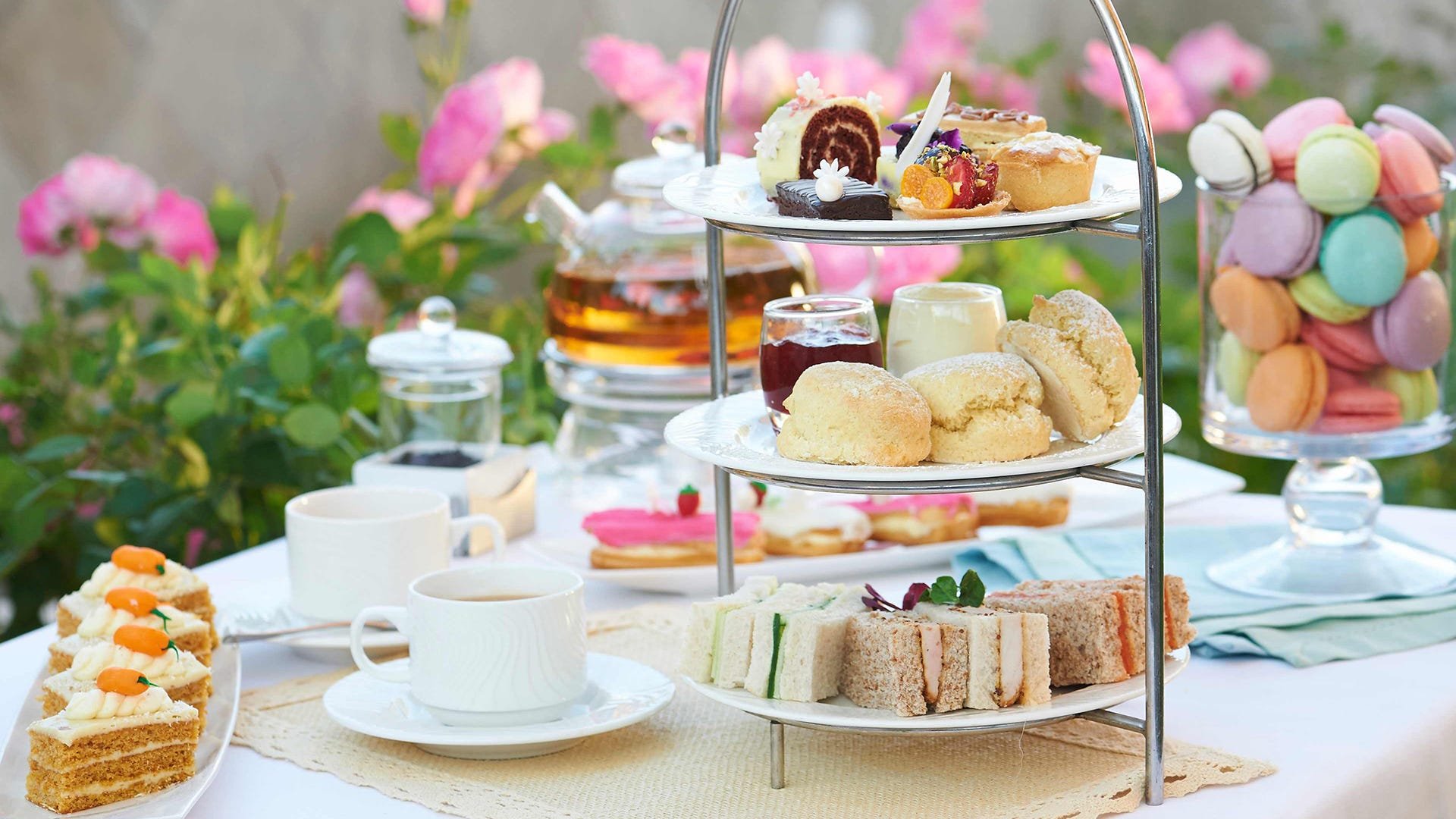 A tiered tea party stand with assorted cakes, cookies, and finger sandwiches, surrounded by teacups, teapot, and a jar of colorful macarons, set outdoors with pink flowers in the background.