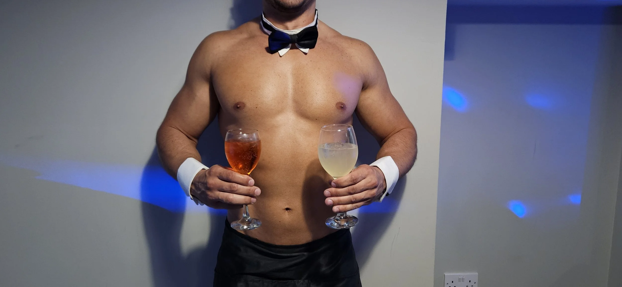 A shirtless man wearing a bow tie and cuffs, holding two glasses of wine, standing against a wall with blue light spots.