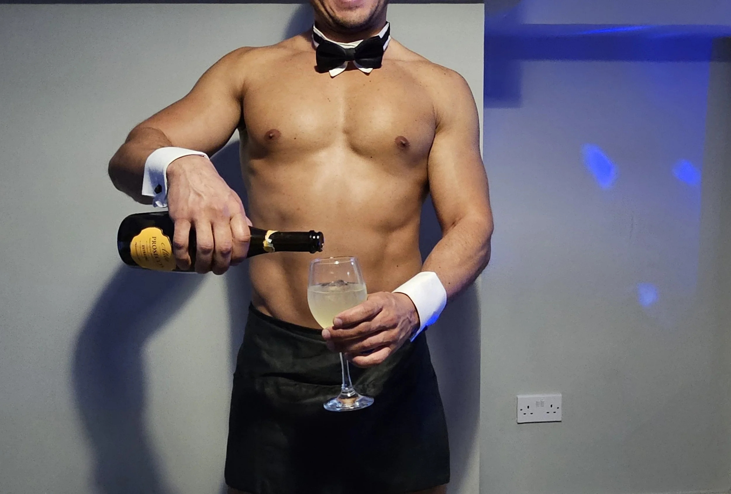 A shirtless man wearing a black bow tie and cufflinks, holding a bottle of champagne, and a wine glass in his other hand, standing against a plain wall with a blue light projection and an electrical outlet visible.