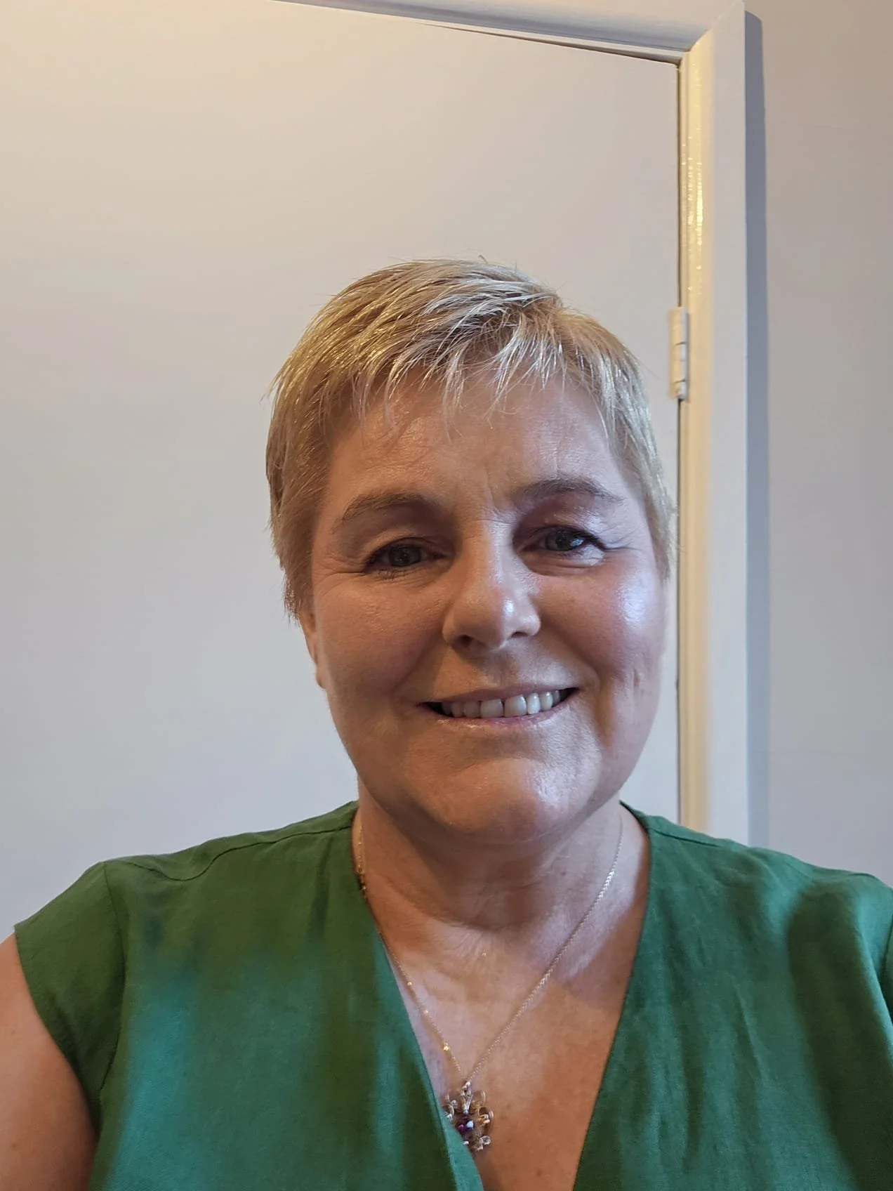 A smiling middle-aged woman with short blonde hair wearing a green shirt and a gold necklace, standing in front of a plain white wall and a doorframe.
