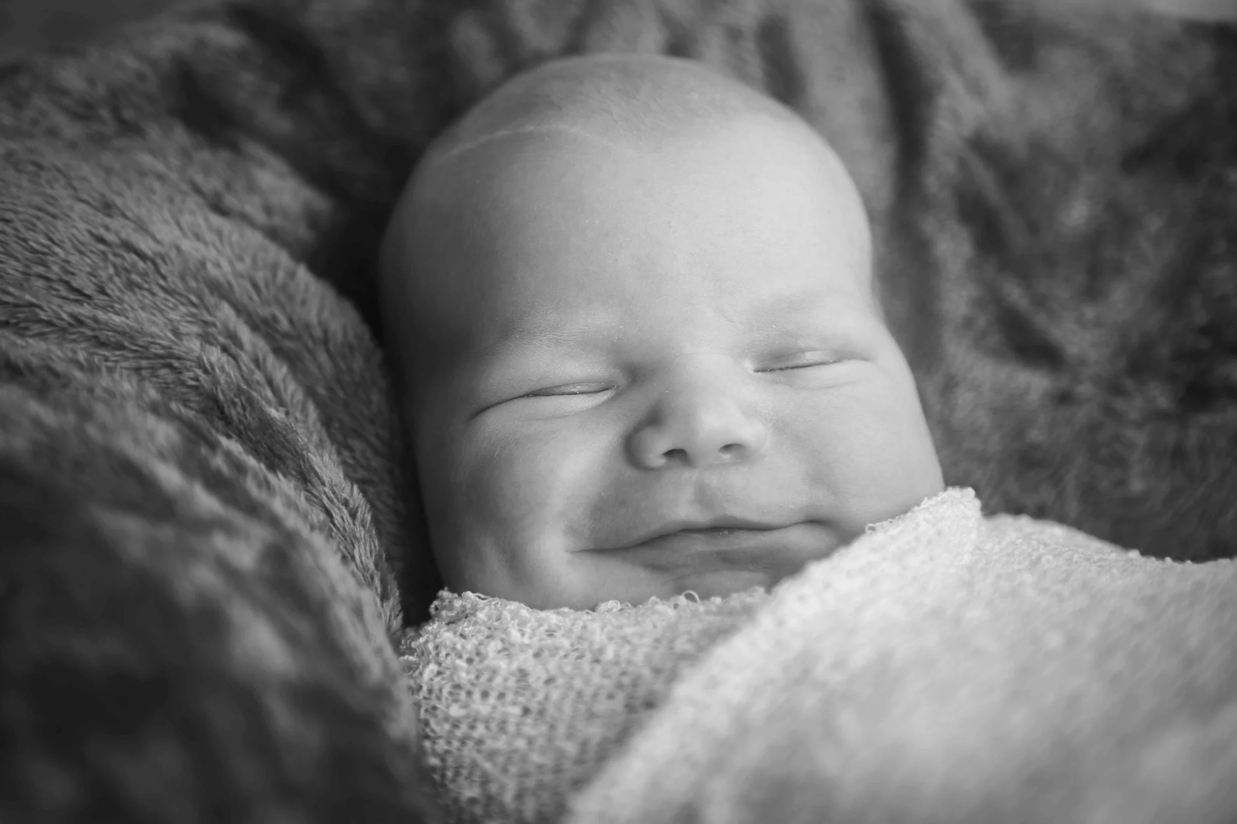 A close-up black and white photo of a smiling newborn baby sleeping, wrapped in a soft blanket.