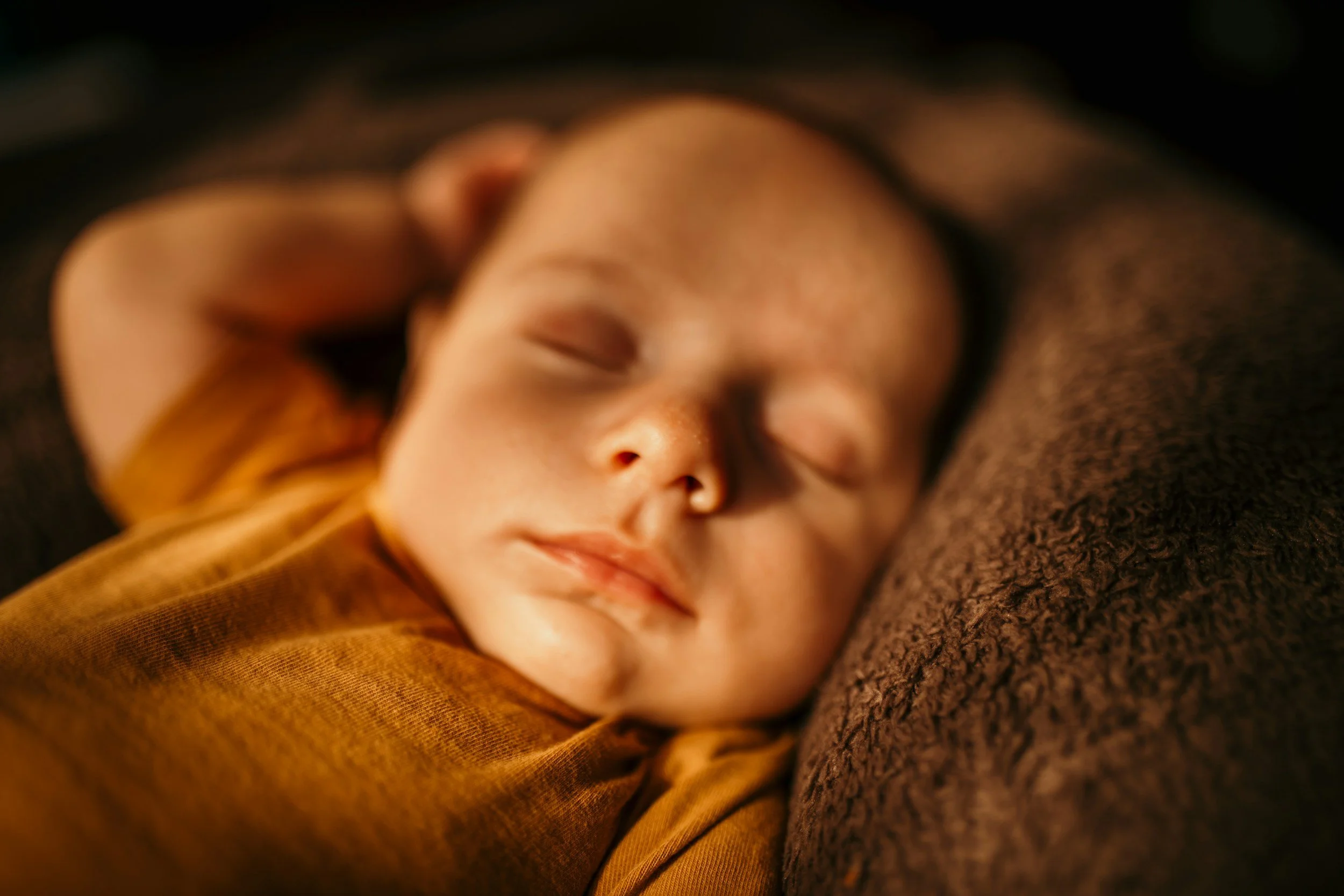 Close-up of a sleeping baby lying on a brown textured pillow, wearing a mustard-colored shirt.
