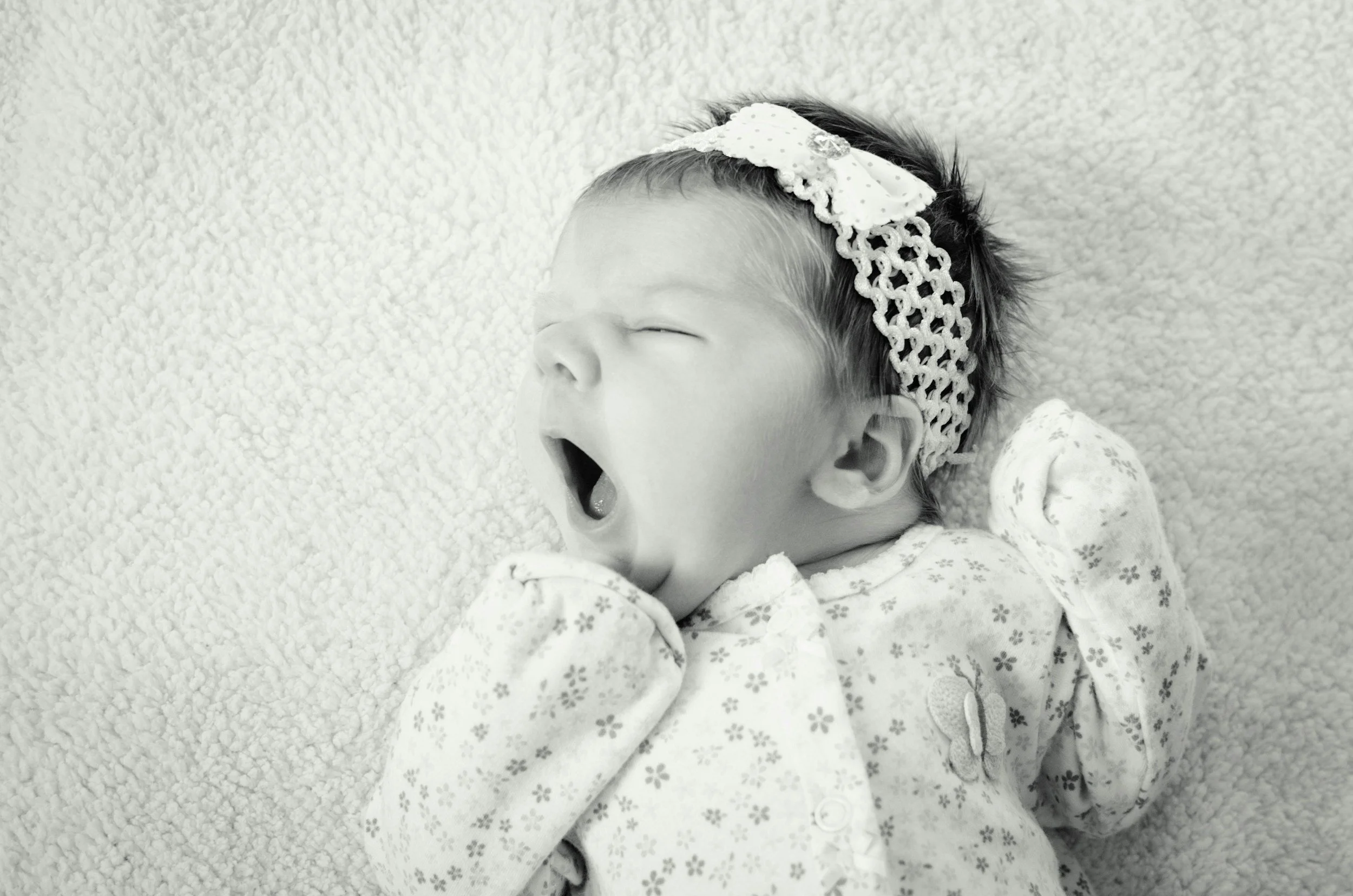 A sleeping baby yawning on a soft textured surface, wearing a floral onesie and a headband with a bow.