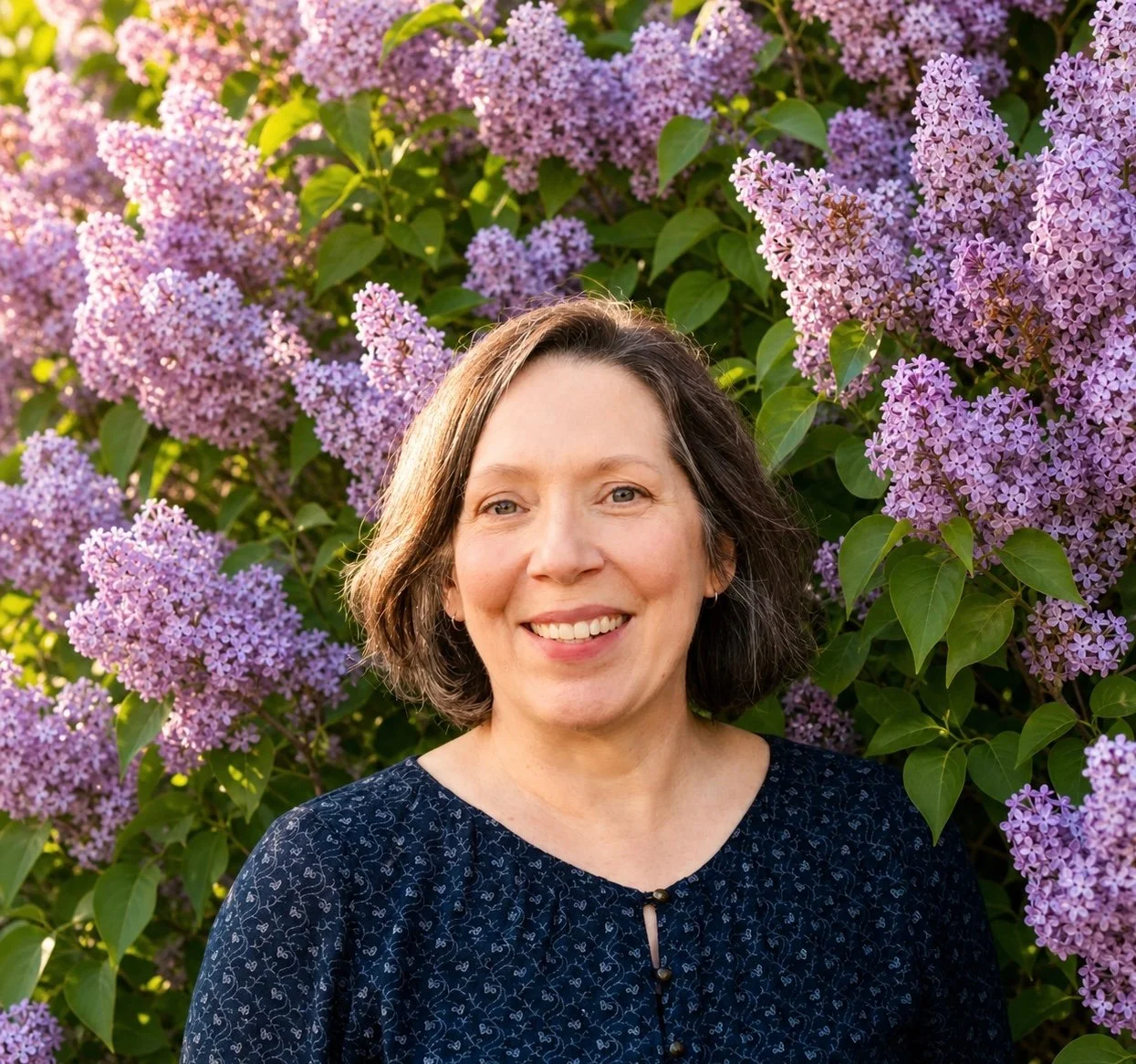 A woman with shoulder-length brown hair smiling in front of a large bush of purple lilac flowers.
