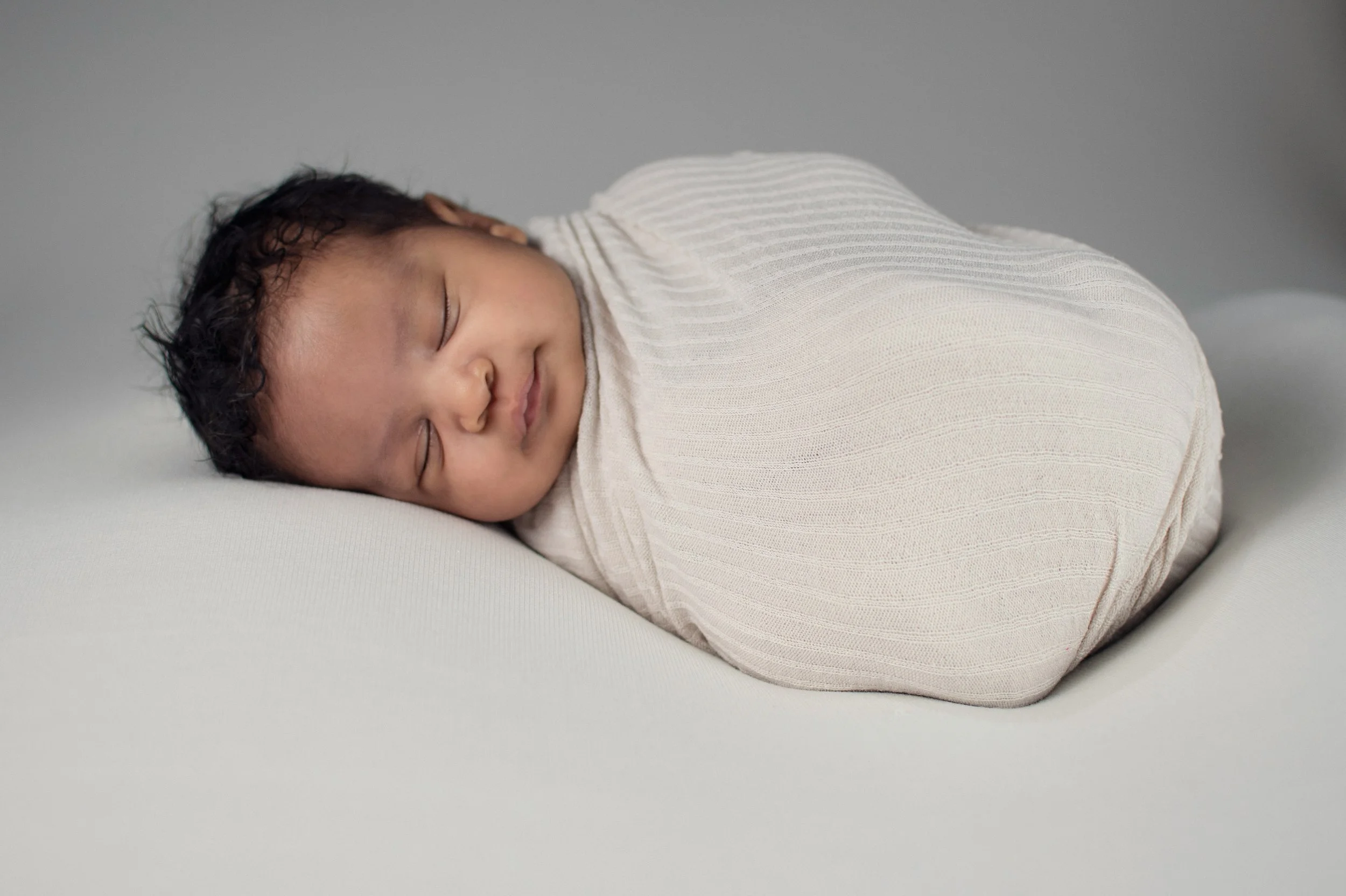 A sleeping baby curled up on a white surface, wrapped in a light-colored, ribbed blanket.