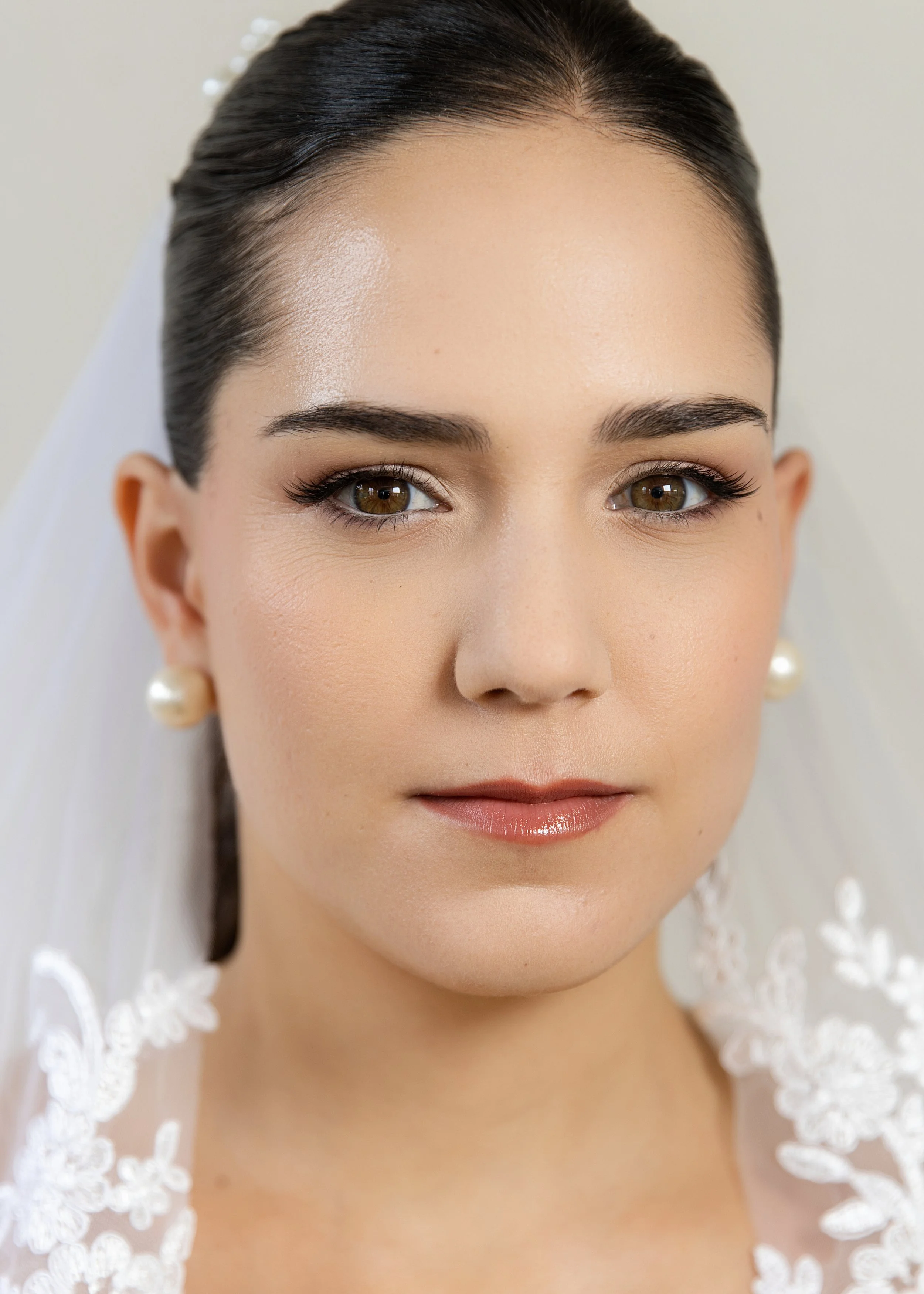 Close-up portrait of a woman with dark hair, brown eyes, wearing pearl earrings, and a white lace veil, possibly a bride.