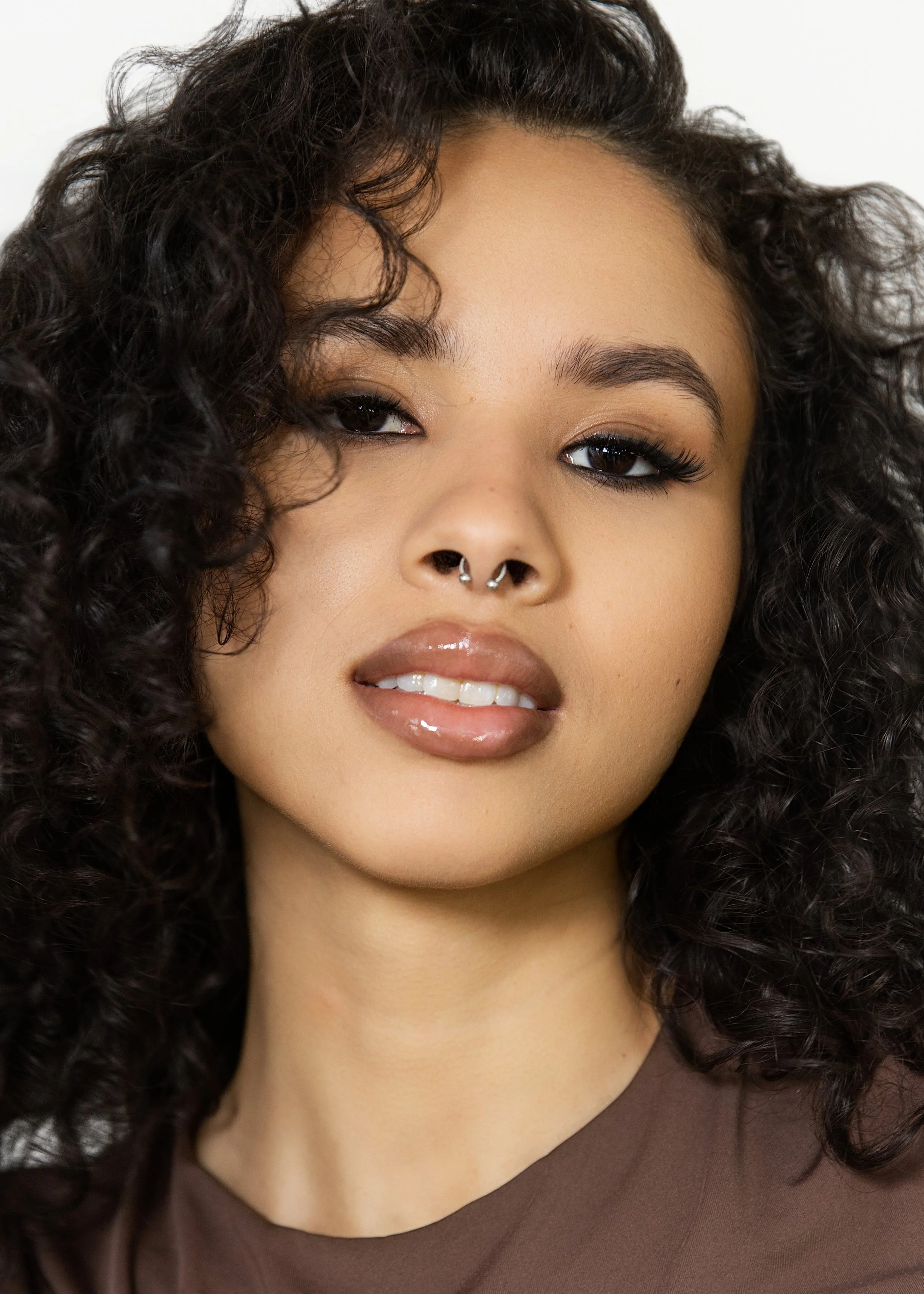 Close-up of a young woman with dark curly hair and a septum piercing, wearing glossy lipstick and a brown top.
