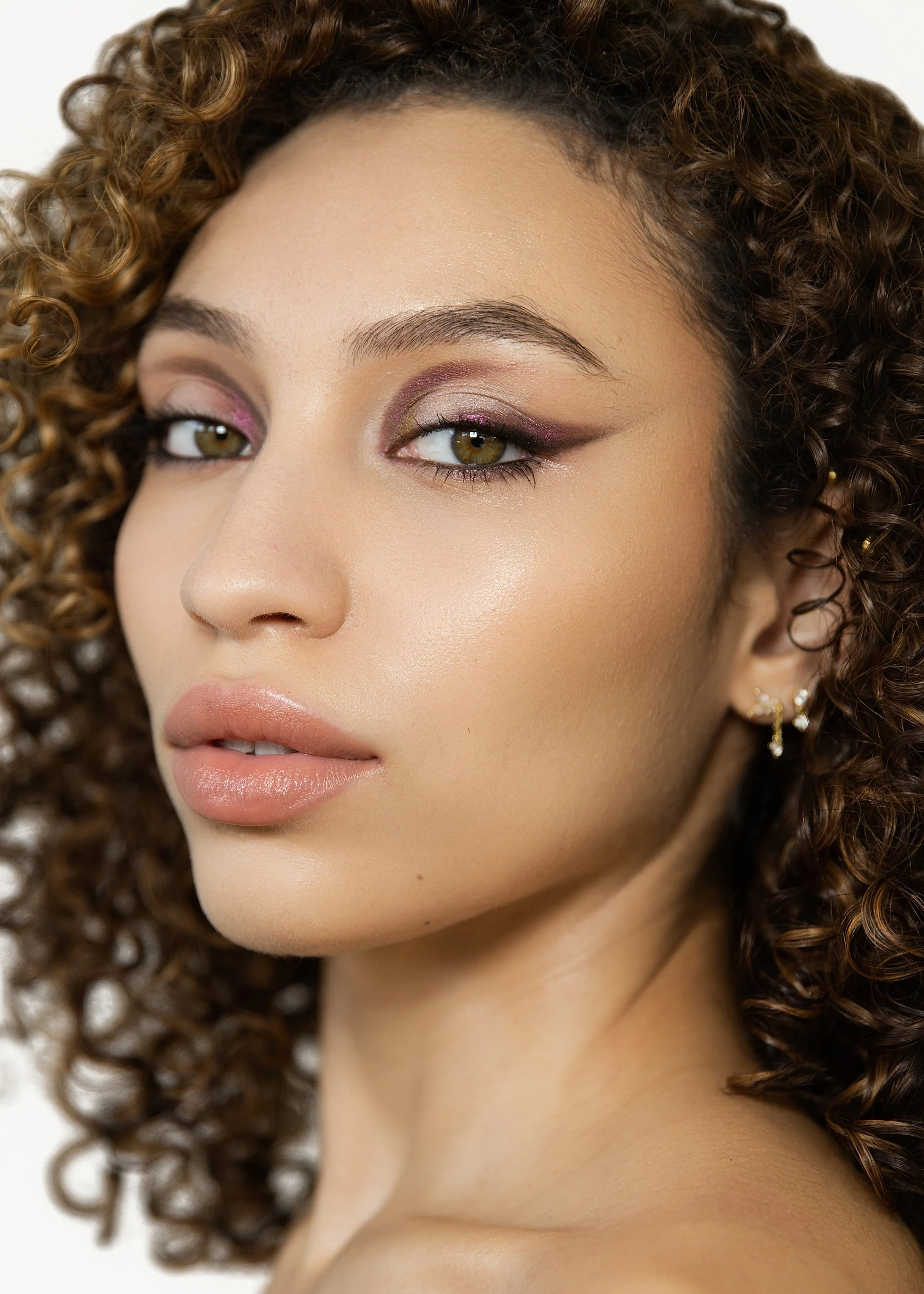 A close-up of a young woman with brown curly hair, hazel eyes, licensed makeup, and wearing earrings.