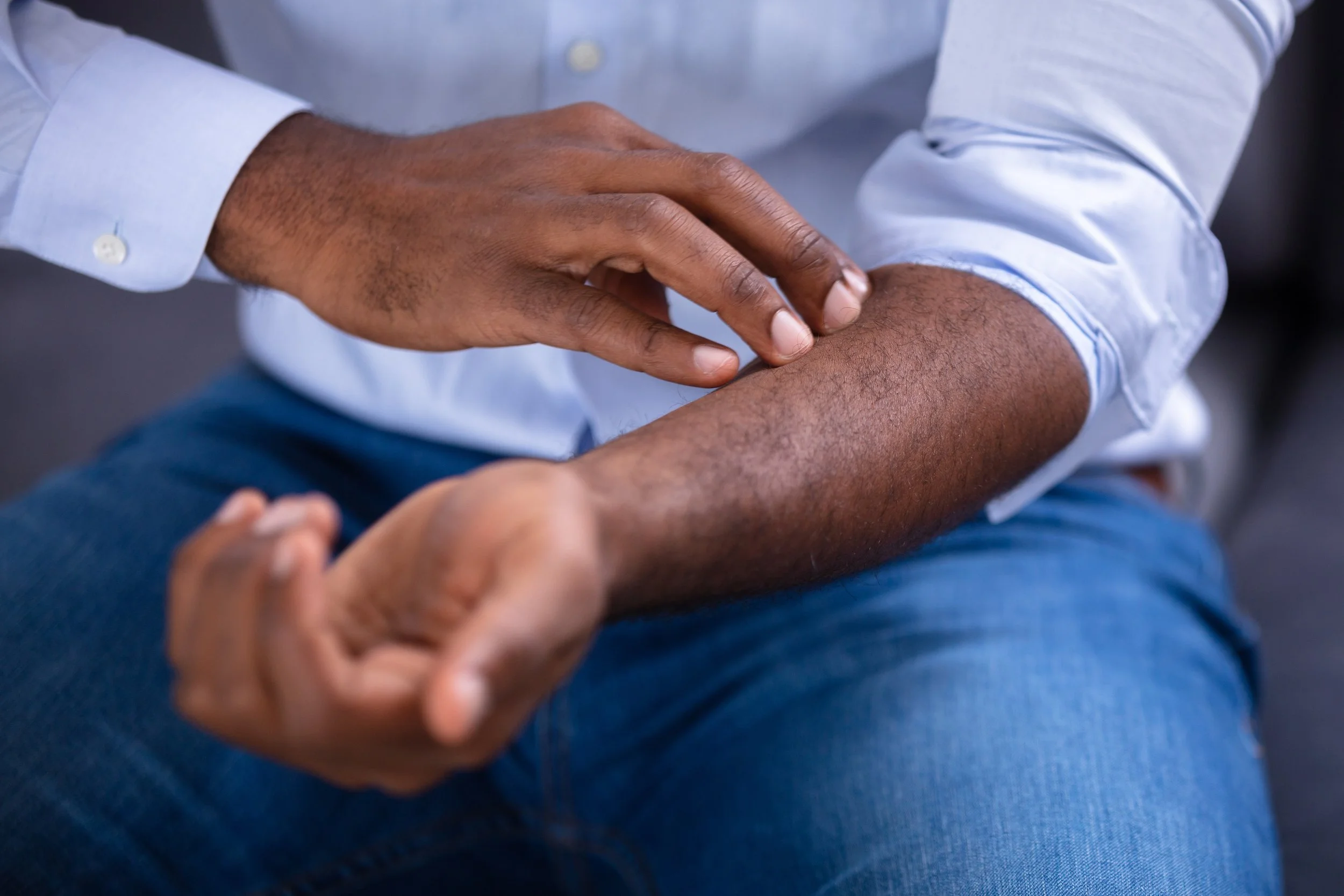 A person with brown skin and short dark hair wearing a light blue dress shirt and dark blue jeans, touching their forearm with their hand.