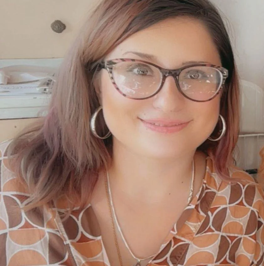 A woman with brown and purple hair wearing glasses, hoop earrings, and a patterned blouse smiling indoors.