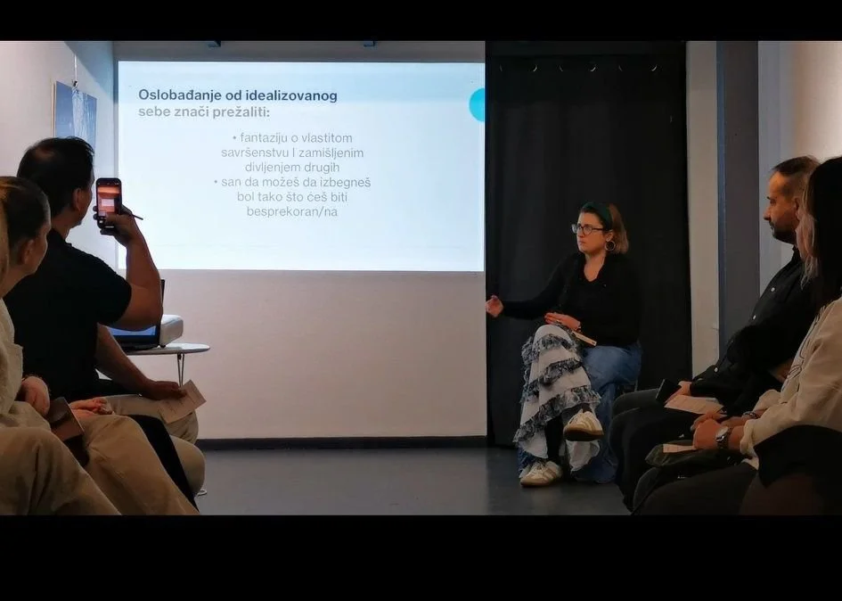 A woman is giving a presentation in a small room with a projector screen, surrounded by an audience. Some audience members are taking photos or notes. The presentation slide has text in Serbian about overcoming idealized self.