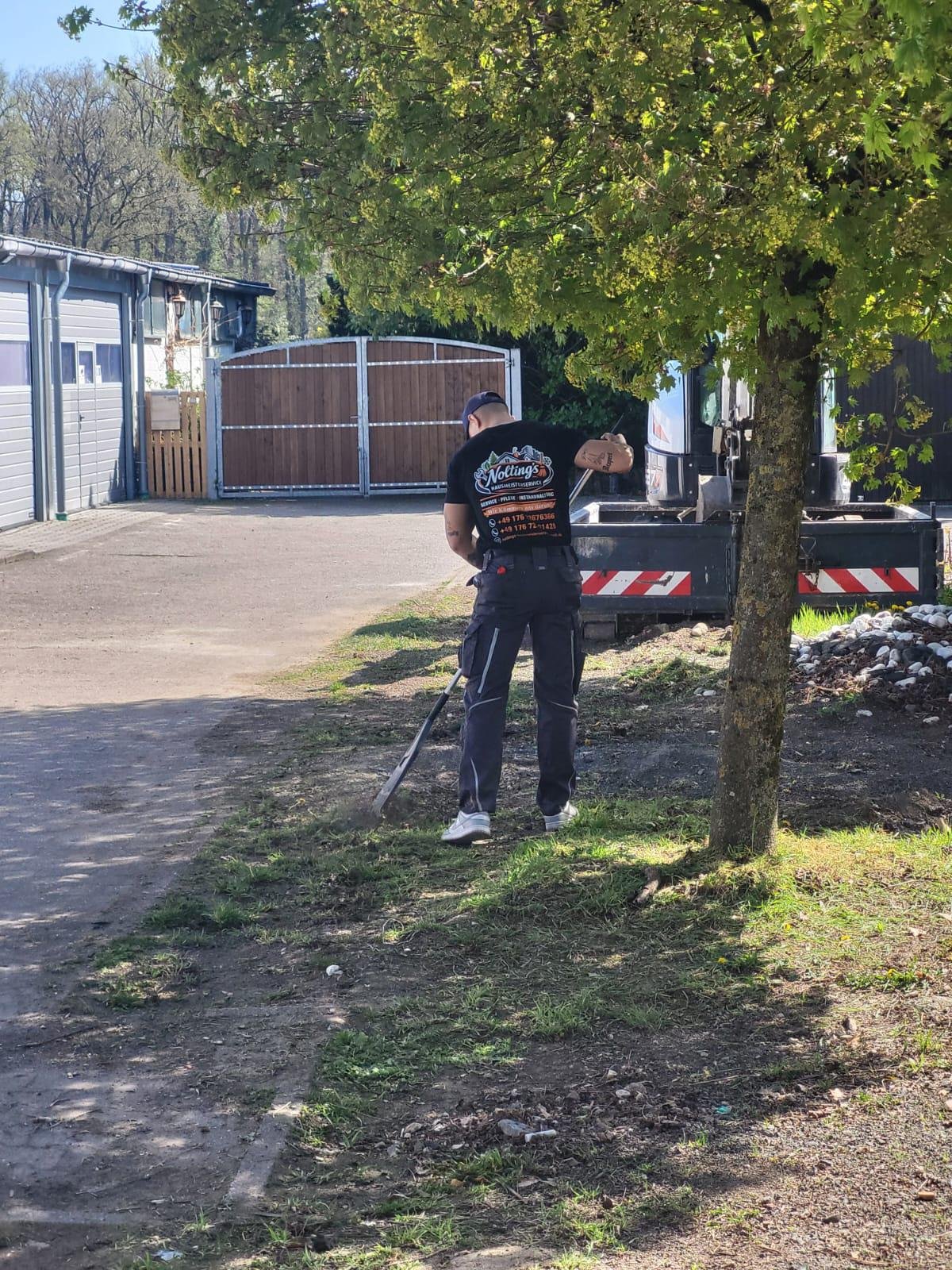 Mitarbeiter versucht den Boden zu ebnen oder zu reinigen, während er mit einem Rasenmäher arbeitet, neben einem Baum und vor einem großen schwarzen Fahrzeug im Freien