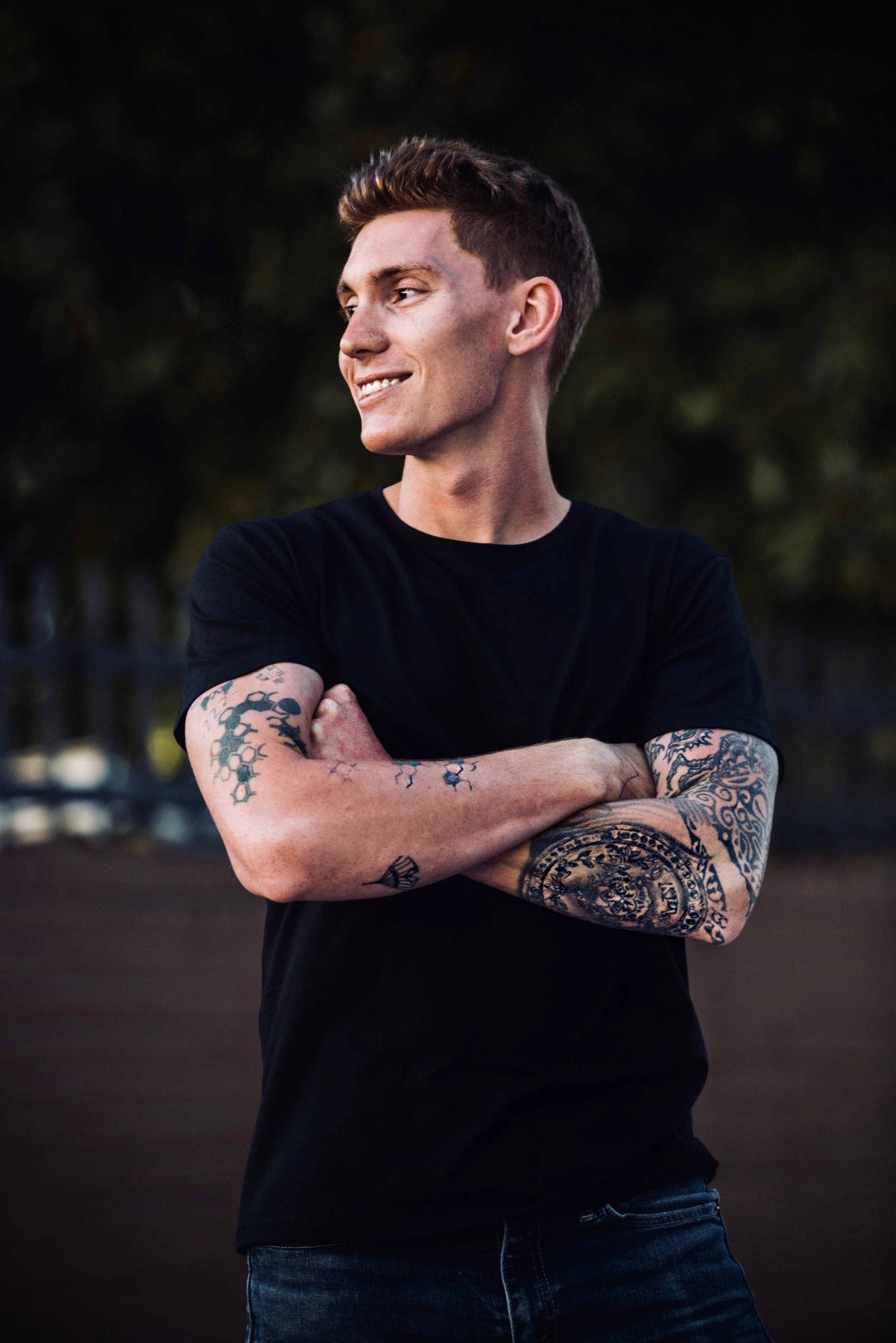 A young man with short brown hair, wearing a black t-shirt and jeans, standing outdoors with his arms crossed, smiling slightly, with tattoos on his arms and a dark blurred background.