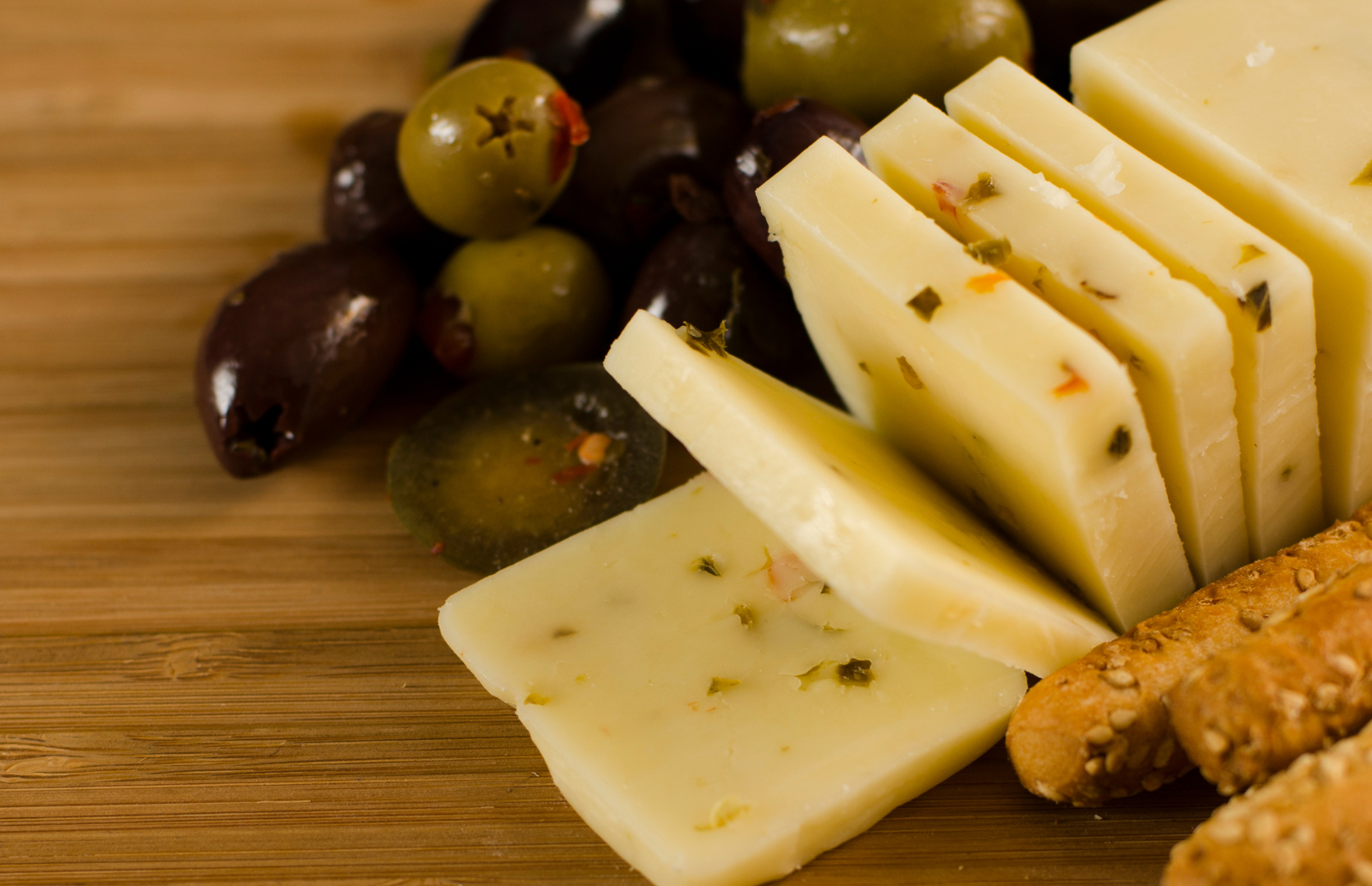 A cheese platter with slices of herb cheese, green and black olives, and breadsticks on a wooden board.