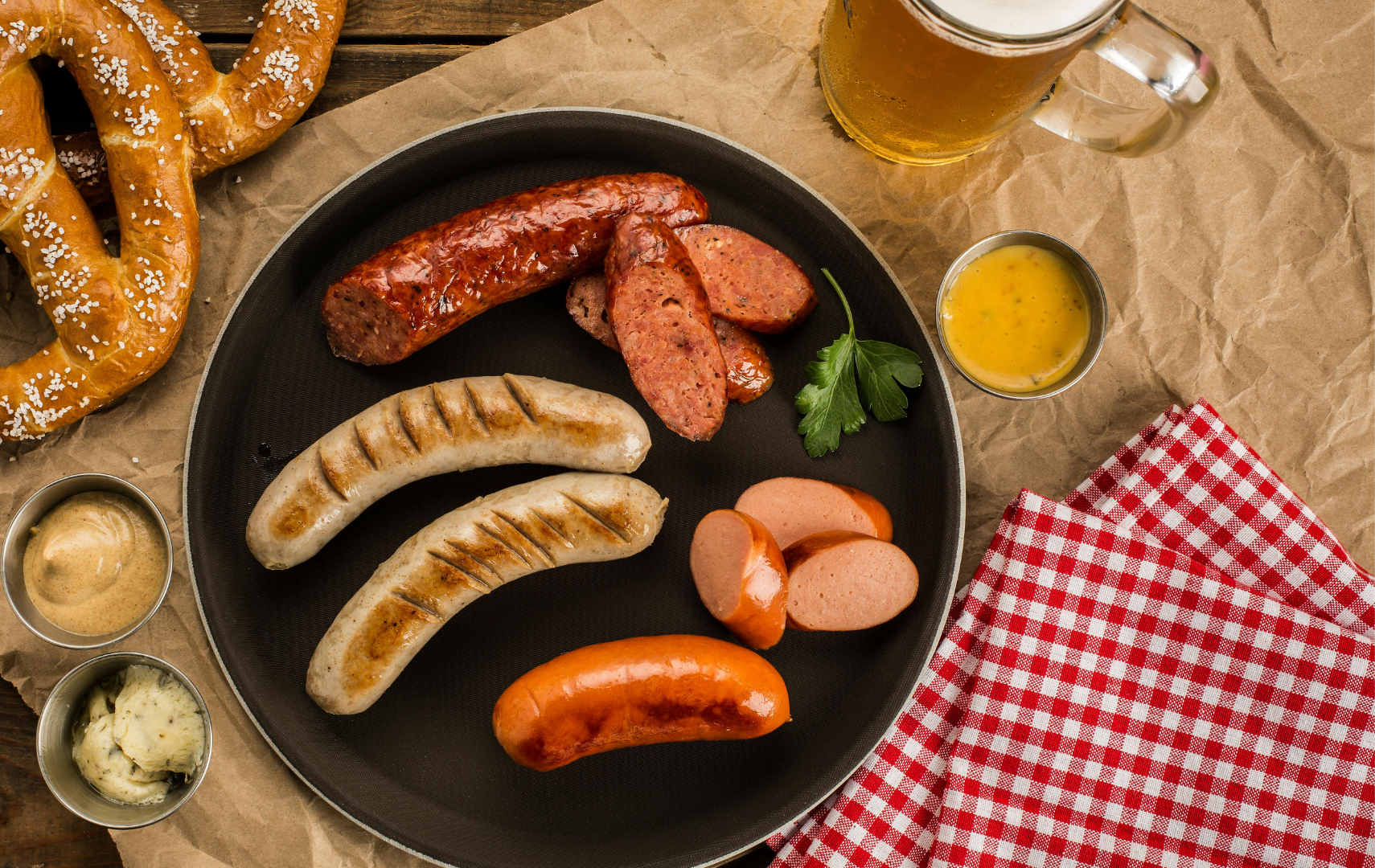 A plate of assorted grilled sausages, sliced salami, and a sausage link with mustard, served with pretzels, a glass of beer, and a cup of beer cheese in a casual setting with red checkered napkins.