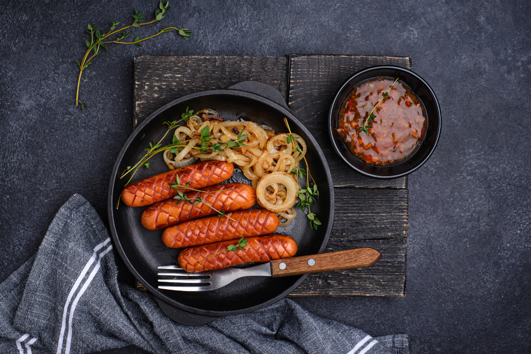 Grilled sausages with sautéed onions and herbs on a black plate, accompanied by a bowl of spicy chili sauce.