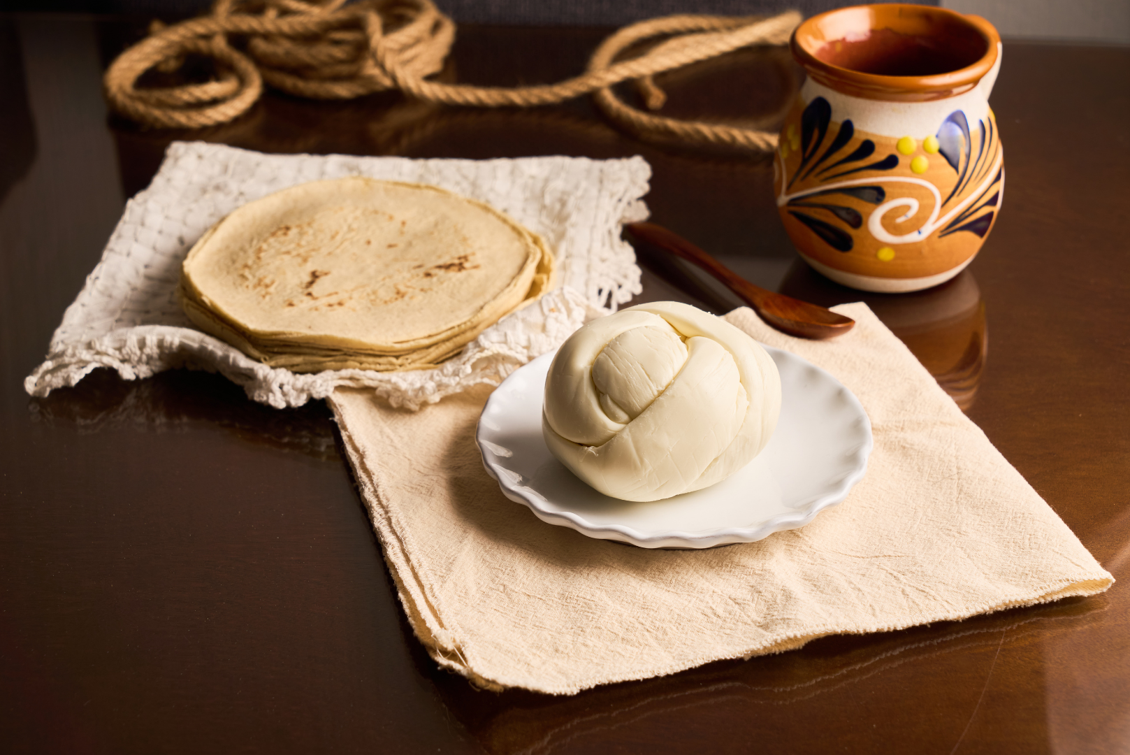 Freshly baked bread rolls, a small white plate with a ball of mozzarella cheese, a woven cloth, a wooden spoon, and an orange and black painted ceramic mug on a dark wooden table.