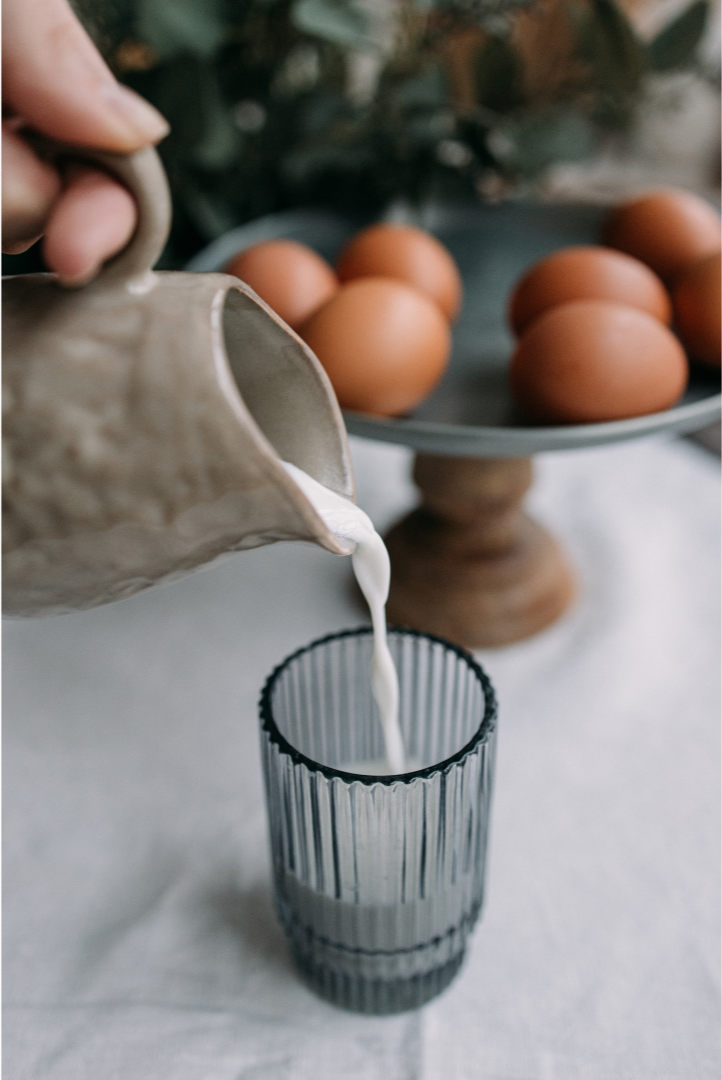 A person pouring milk from a beige ceramic pitcher into a tall, ridged glass. In the background, there is a cake stand with brown eggs on it, and some greenery.