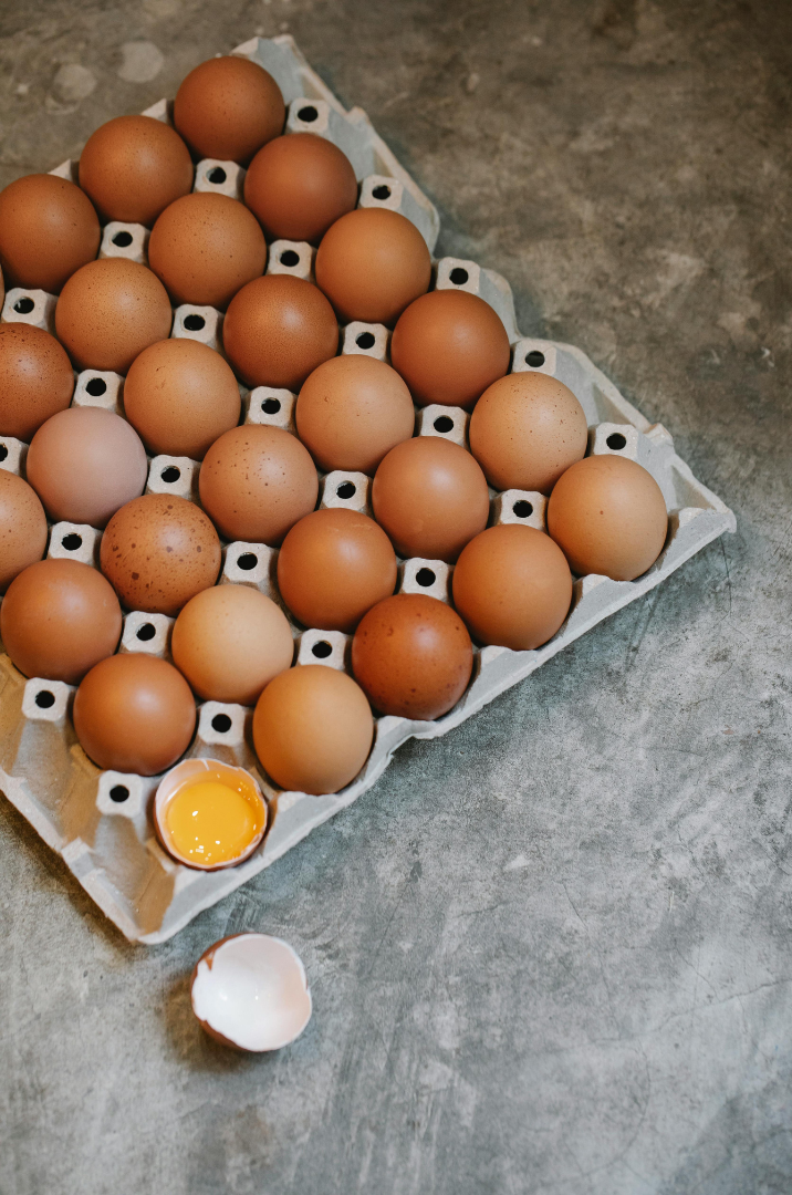 A tray of brown eggs with one cracked open showing the yolk inside and an empty eggshell in front on a gray surface.