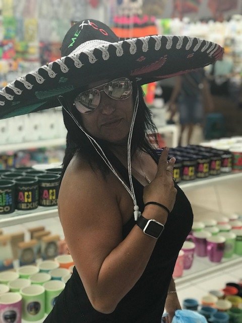 A woman wearing a large sombrero, sunglasses, and a black tank top, pointing at her watch in a store aisle with colorful mugs and containers on shelves.