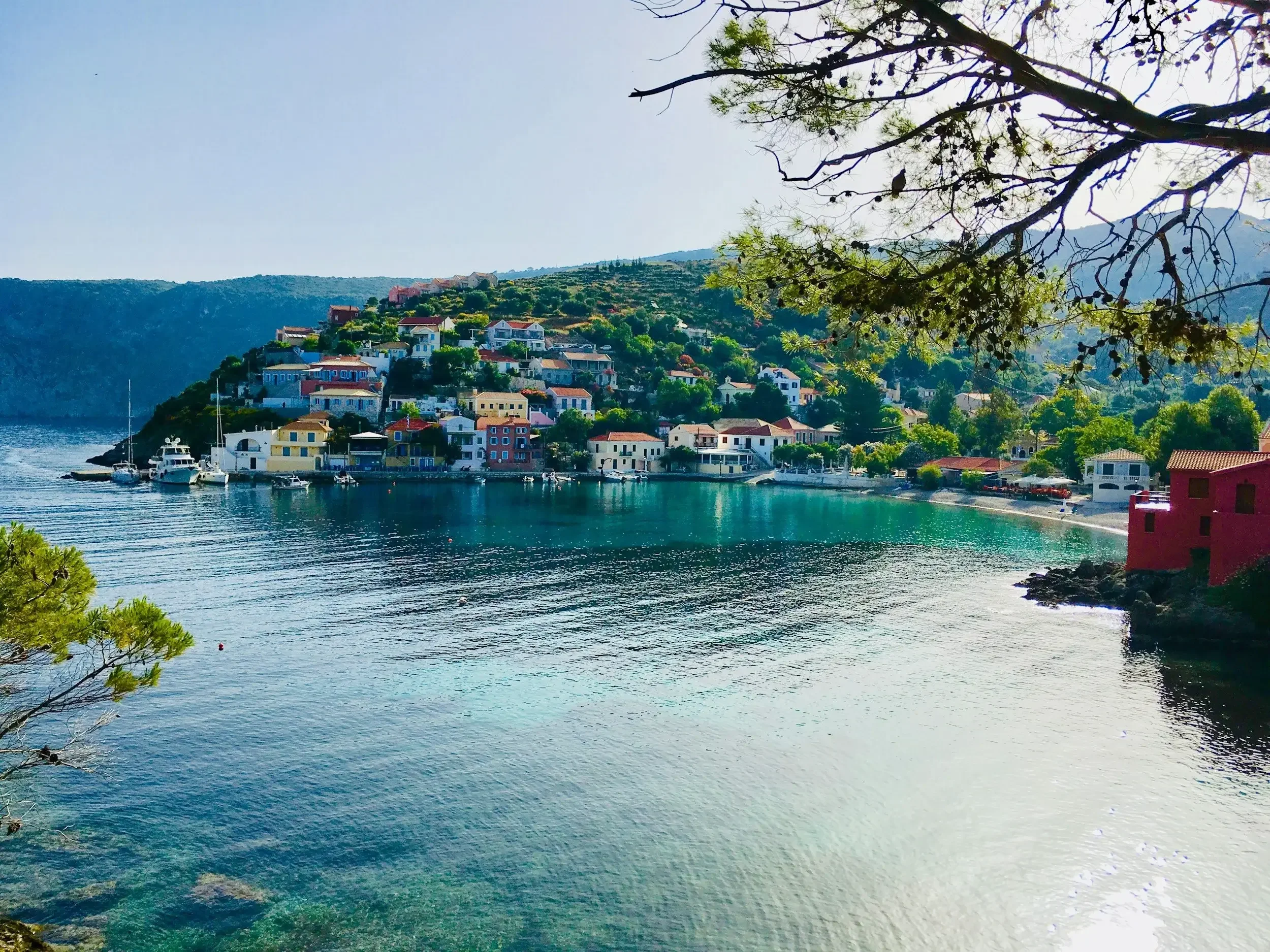 A coastal village with colorful houses on a hill overlooking a calm bay with boats. Trees frame the scene with a bright sky and mountains in the background.