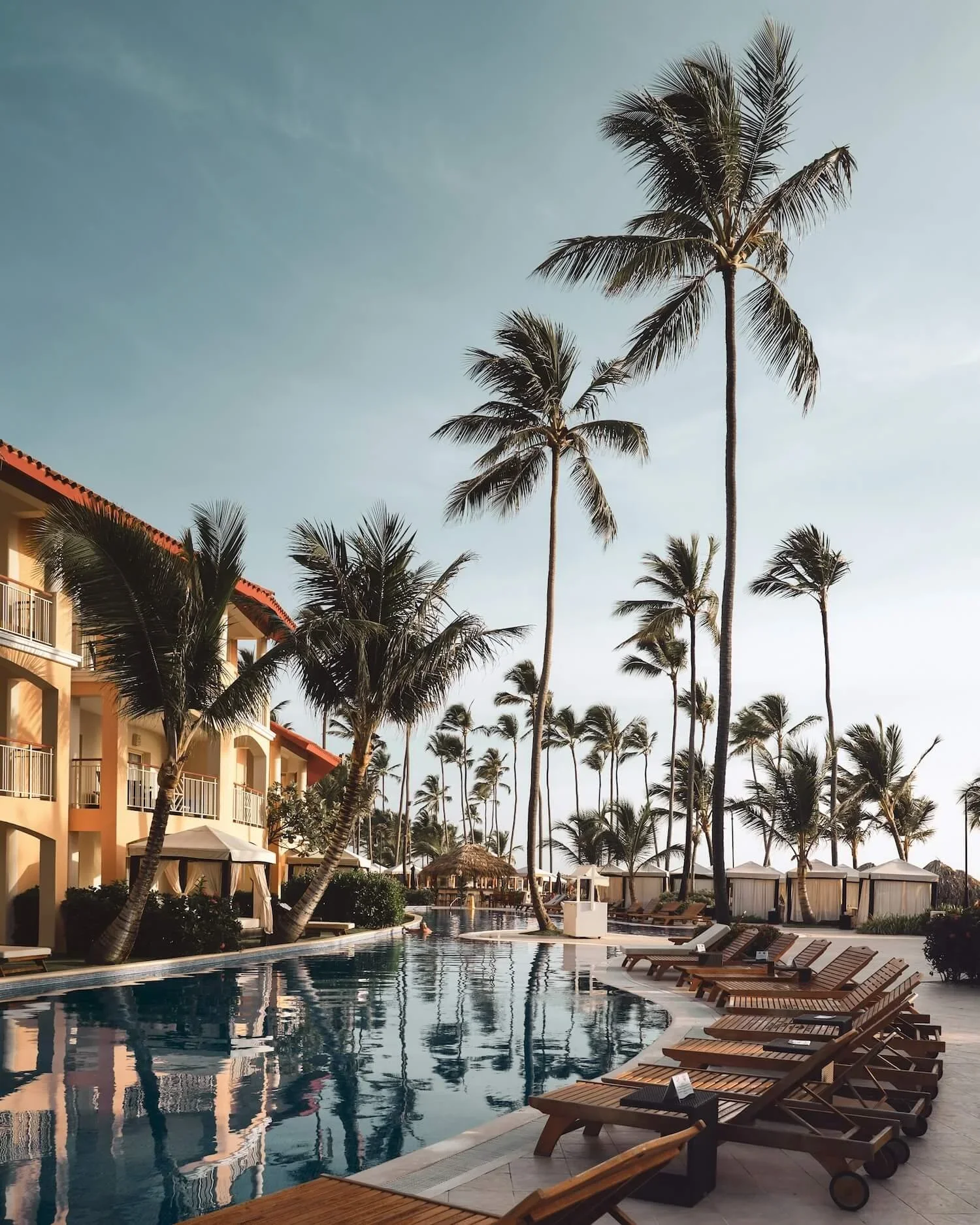 A resort swimming pool with lounge chairs and tall palm trees under a slightly cloudy sky.