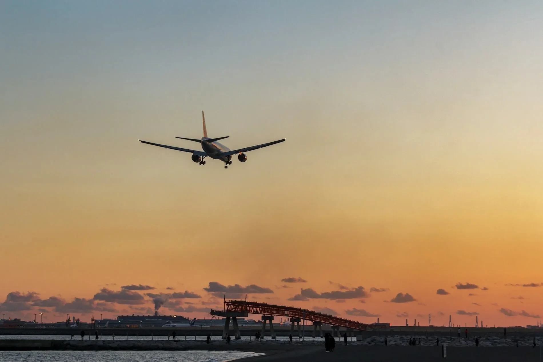 An airplane approaching to land at an airport during sunset, with a pier and people along the waterfront in the foreground.
