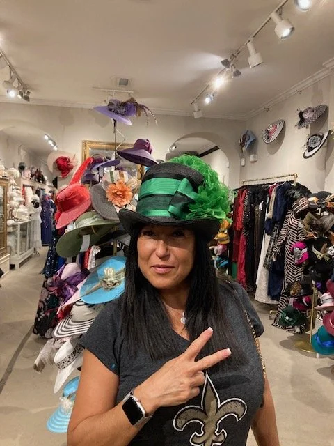 Woman trying on a large, green and black striped hat with a feather and flowers in a hat shop, surrounded by various colorful hats.