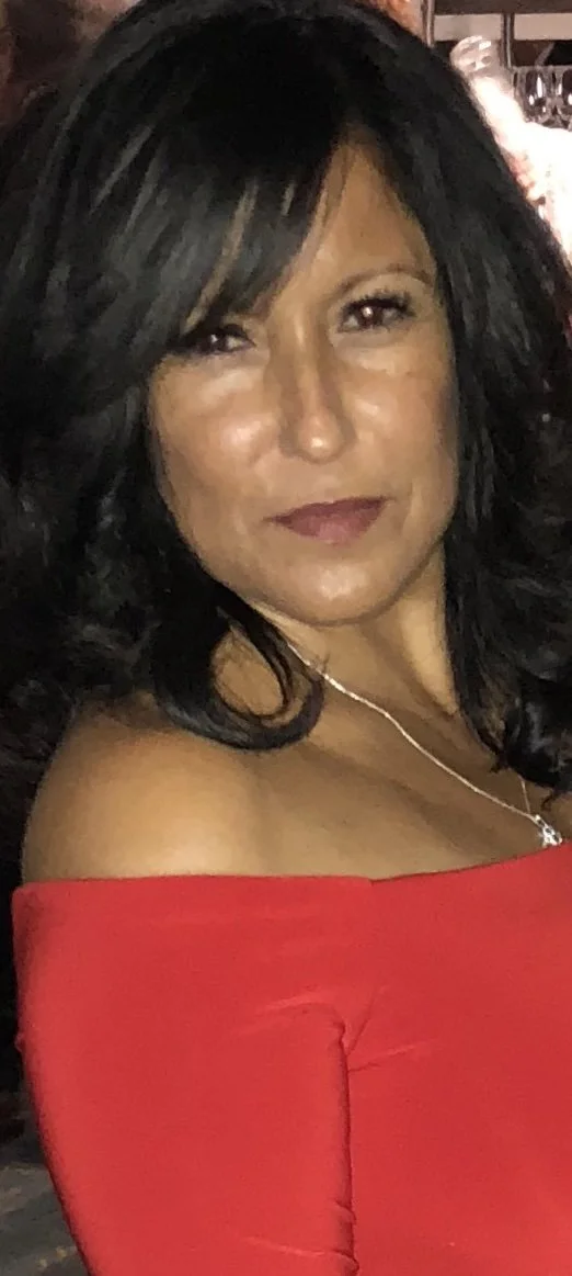 A woman with black hair styled in loose waves, wearing a red off-shoulder top and a silver necklace, smiling gently in a close-up portrait.