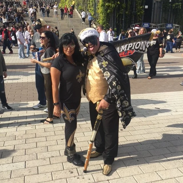 Two people dressed in New Orleans Saints fan attire standing outdoors in a crowded area, with one holding a cane and a flag that says 'Super Bowl Champions' in the background.