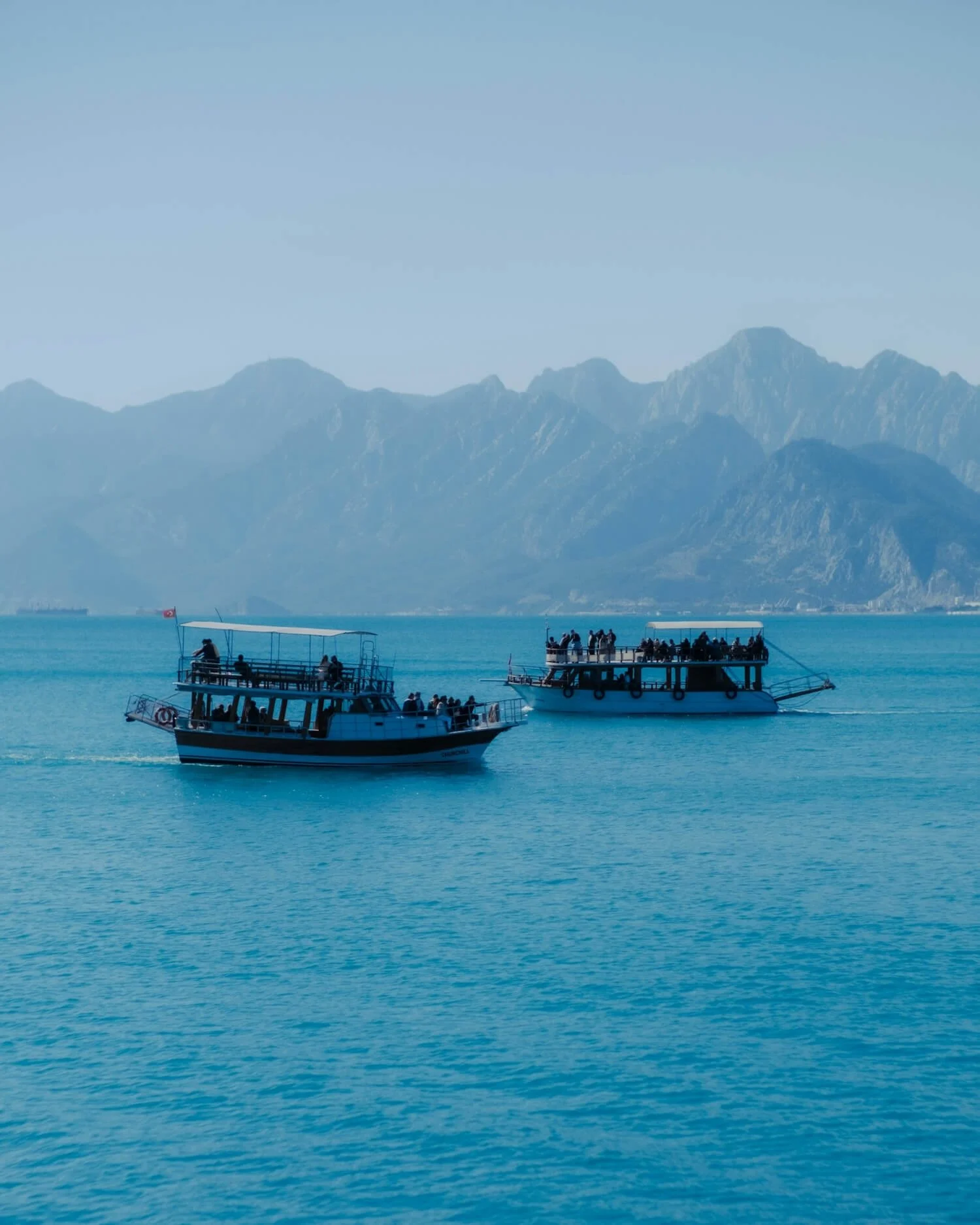 Two boats floating on a calm body of water with mountains in the background, and several people on the boats.