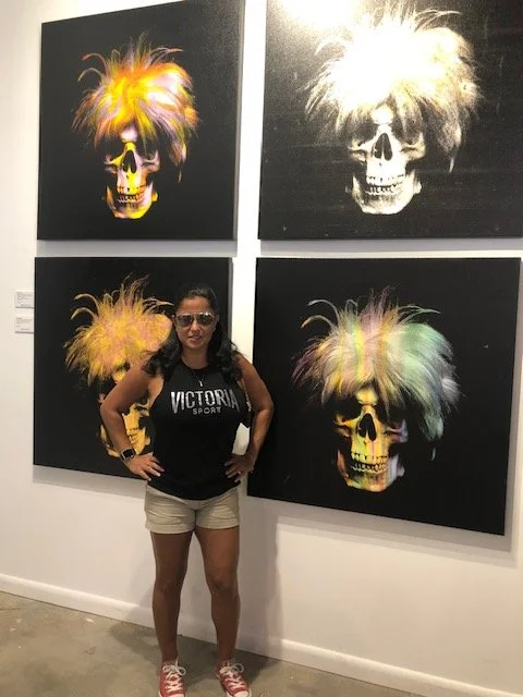 A woman is standing in front of four large colorful art pieces depicting skulls with wild, colorful hair on a black background.
