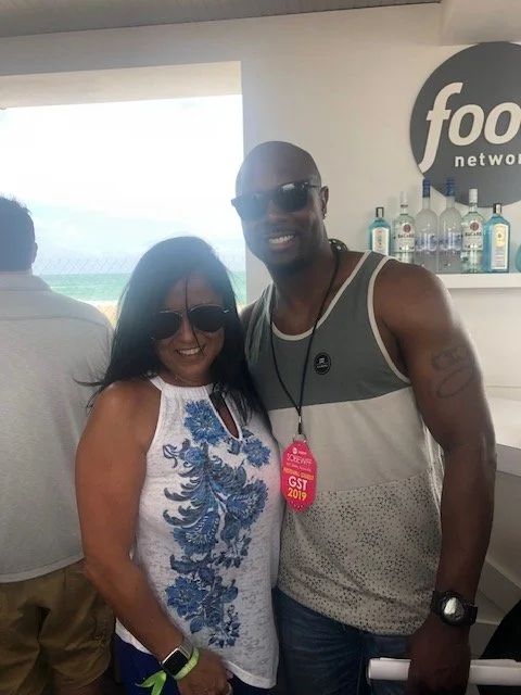 A man and a woman smiling and wearing sunglasses inside a bar or lounge, with bottles on a shelf behind them and a window showing the ocean outside.