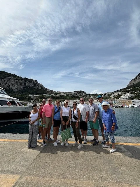 Group of nine people standing on a dock by the water with boats and mountainous coastal scenery in the background.