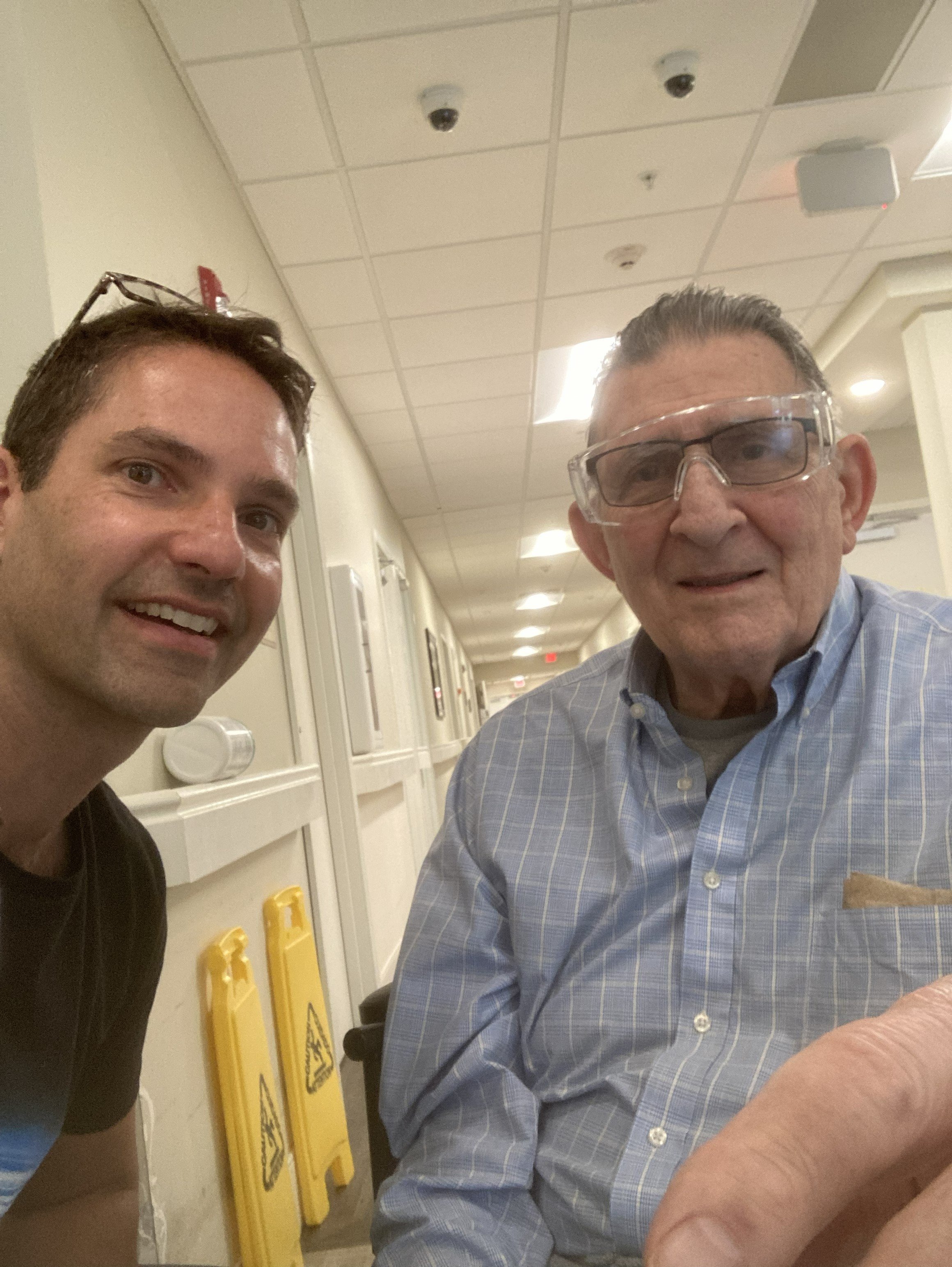 Two men taking a selfie in an indoor setting, possibly a hospital or clinic. One is wearing a black shirt with glasses on his head, and the other is in a blue checkered shirt with protective goggles. Yellow caution signs are visible in the background.