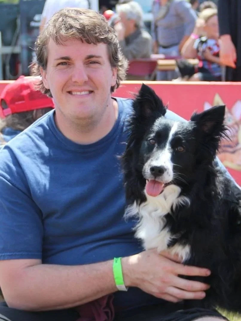 A smiling man in a blue t-shirt sitting outdoors with a black and white Border Collie dog, both looking at the camera.