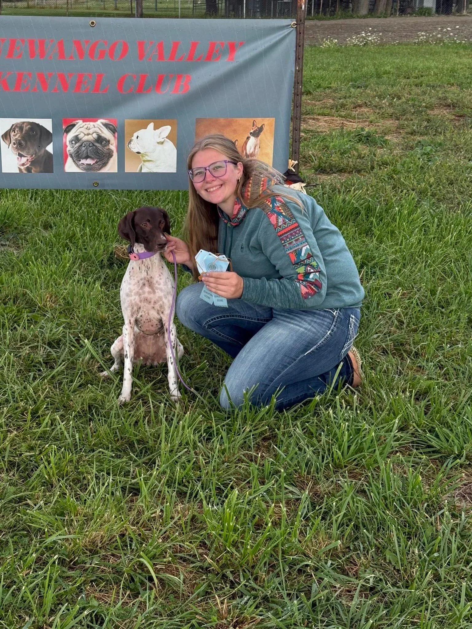 A woman with glasses kneels on grass, smiling, as she holds a certificate and holds the leash of a brown and white dog sitting beside her. Behind them is a banner for the Wewango Valley Kennel Club with pictures of various dogs.