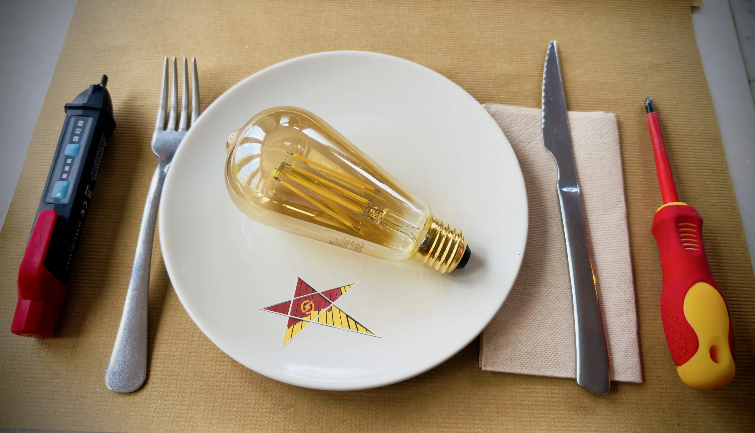 Lamparita de luz dorada sobre plato blanco con logotipo, cubiertos a la izquierda y derecha, sobre manteles beige y de color madera.
