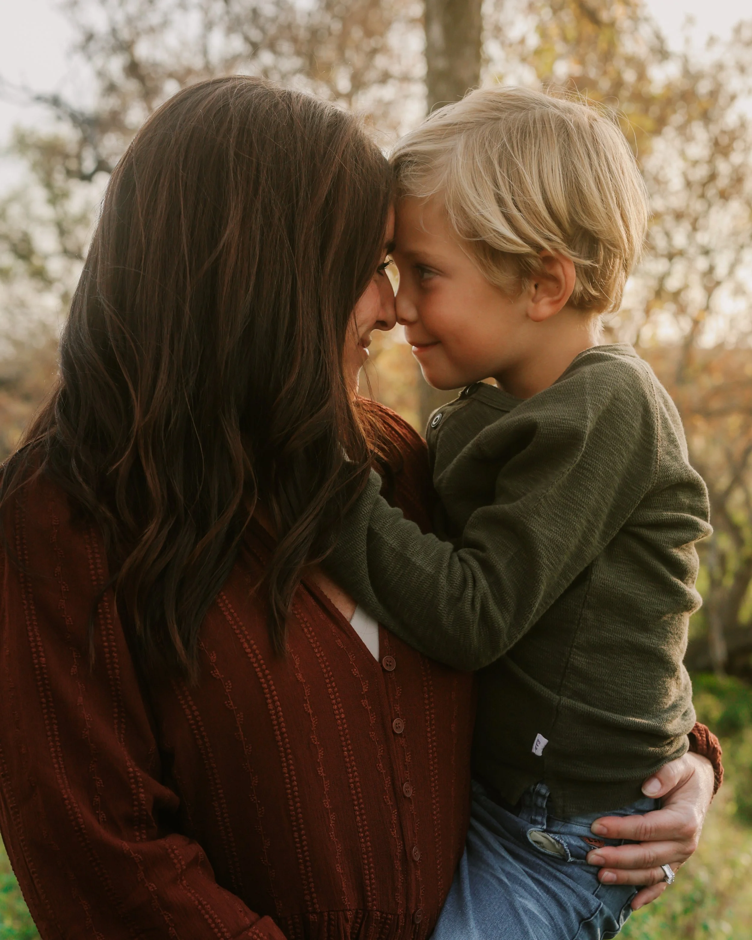 A woman holding a young boy close, their foreheads touching, outdoors during autumn with trees in the background.