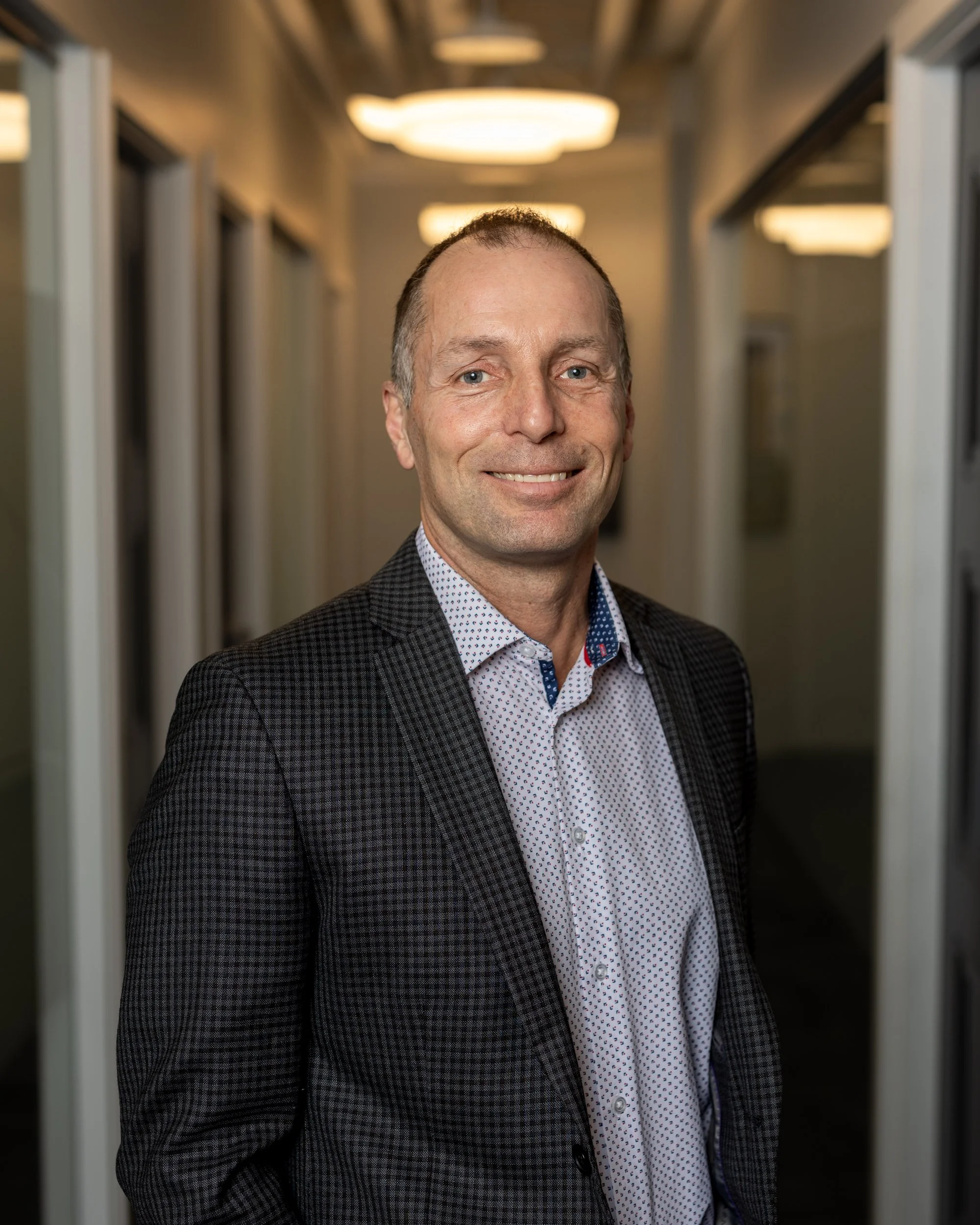 A man in a checked blazer and patterned shirt stands in a corridor with warm lighting. Warm lights are visible on the ceiling behind him.