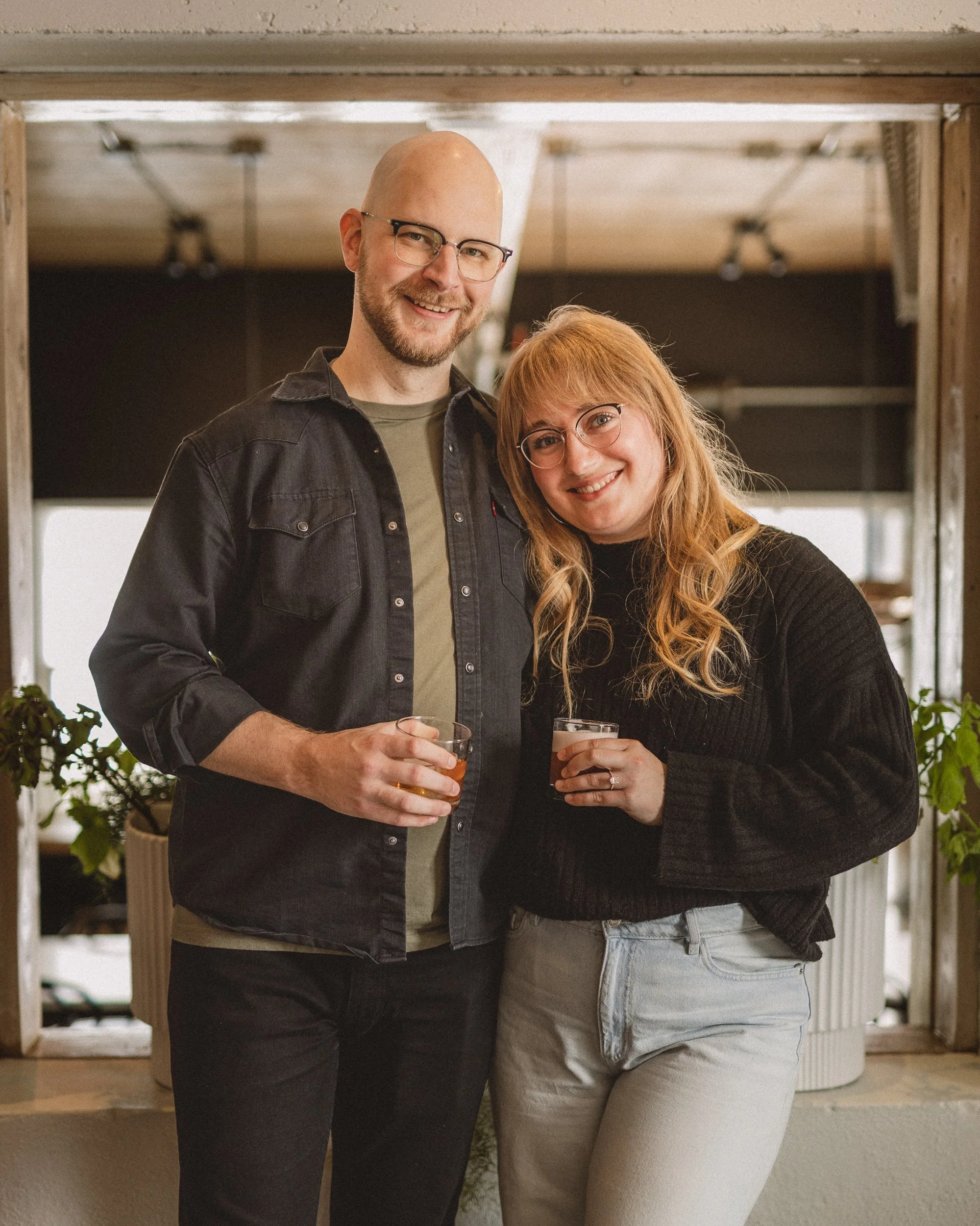 A man and woman smiling, holding drinks, standing close together inside a room with plants and wooden accents.