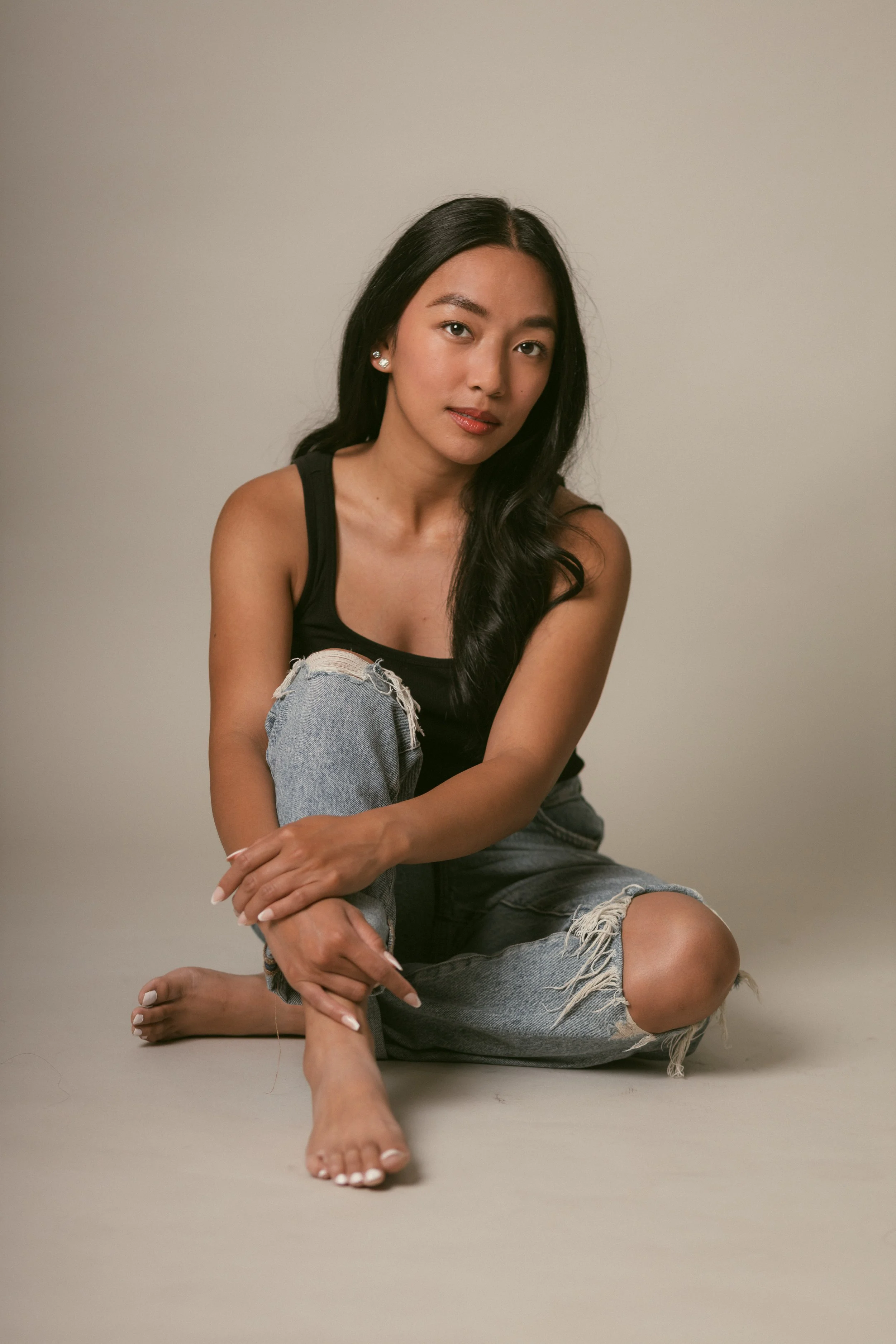 A woman with long black hair, sitting on the floor with one knee up, wearing a black tank top and ripped jeans, looking at the camera against a plain background.